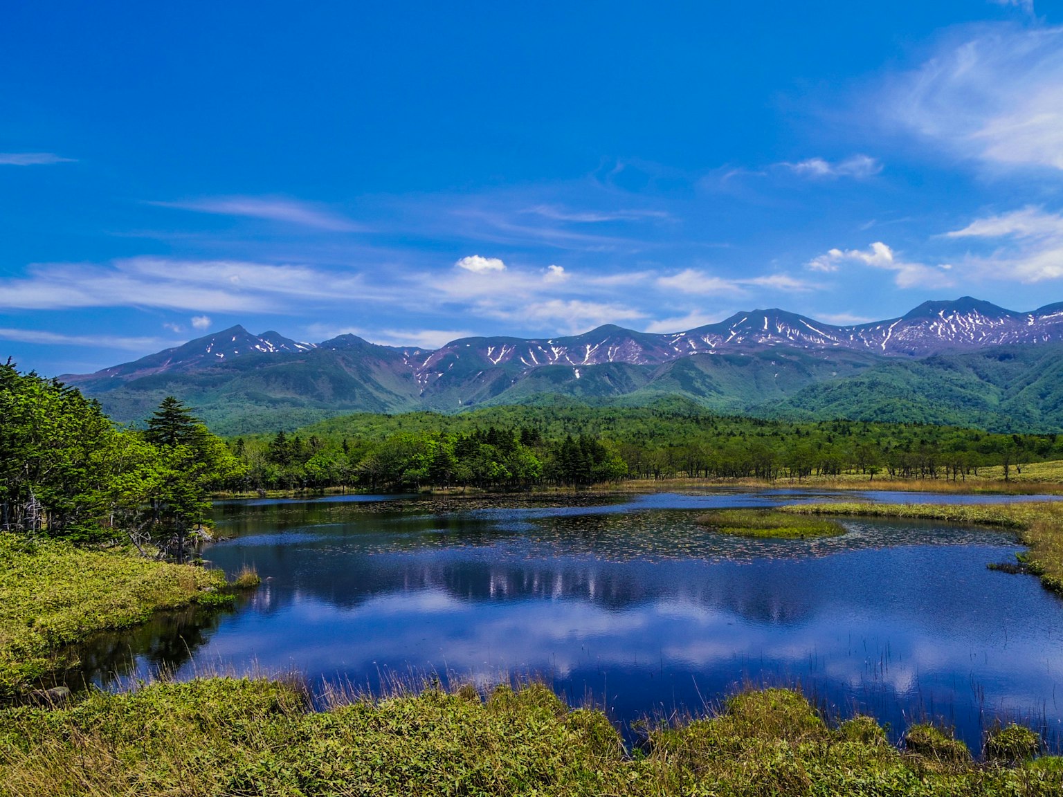 Vista panoramica di un cielo blu chiaro e montagne innevate con un lago tranquillo e prati verdi lussureggianti