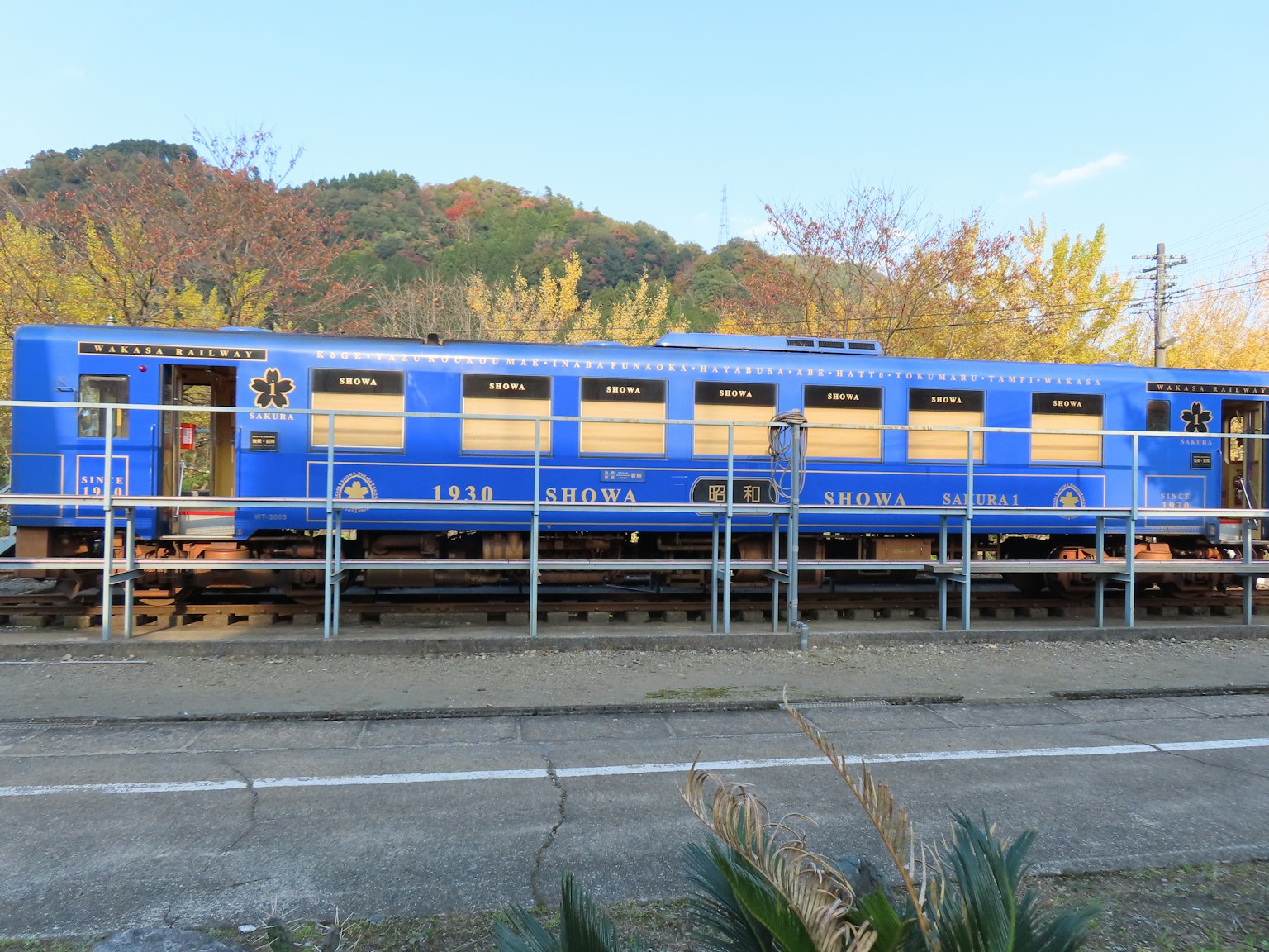 Sebuah kereta biru diparkir di stasiun dikelilingi dedaunan musim gugur