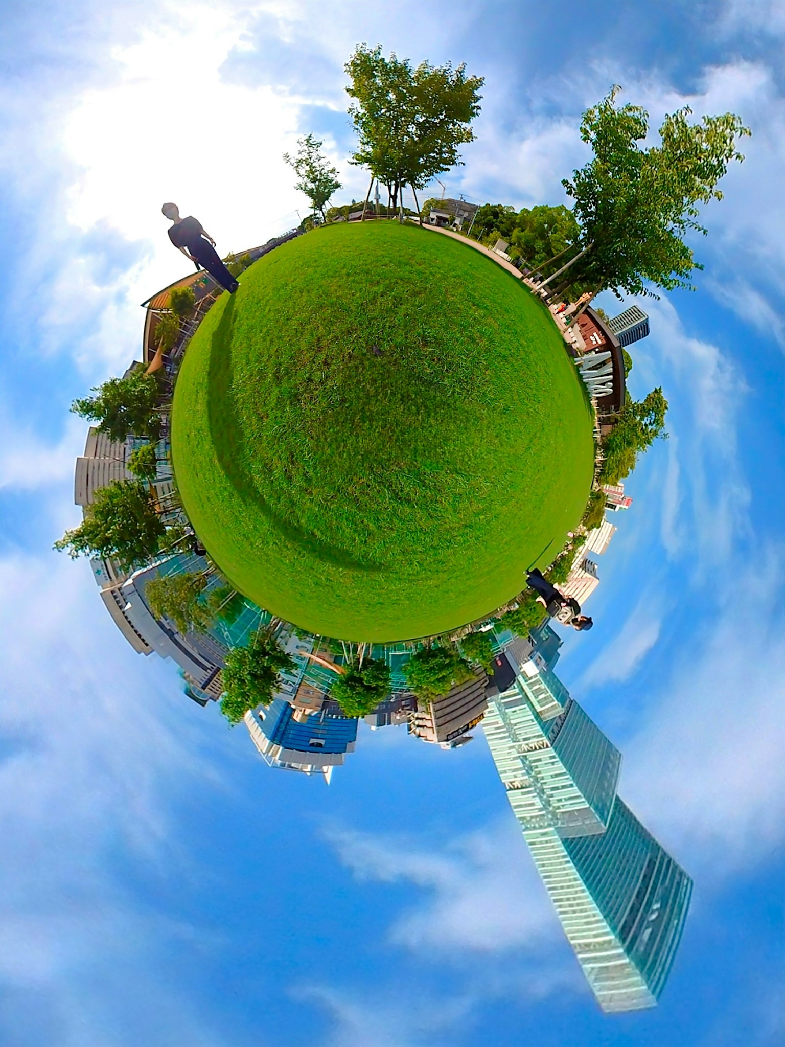 A panoramic image of a small globe with green grass and blue sky featuring surrounding tall buildings and trees