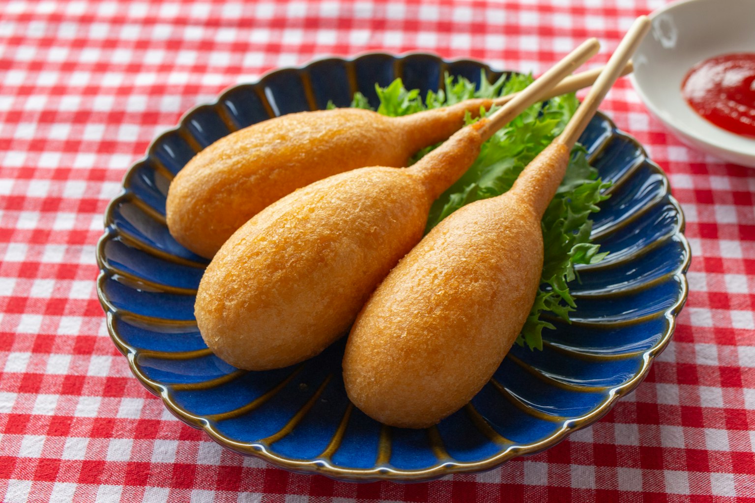 Tiga corn dog di piring biru dengan selada dan saus tomat