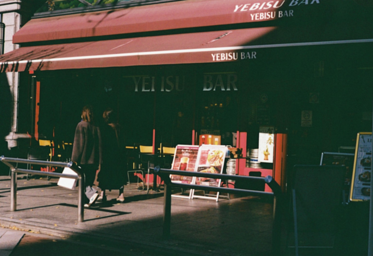 Exterior of a bar with a red awning and people walking by