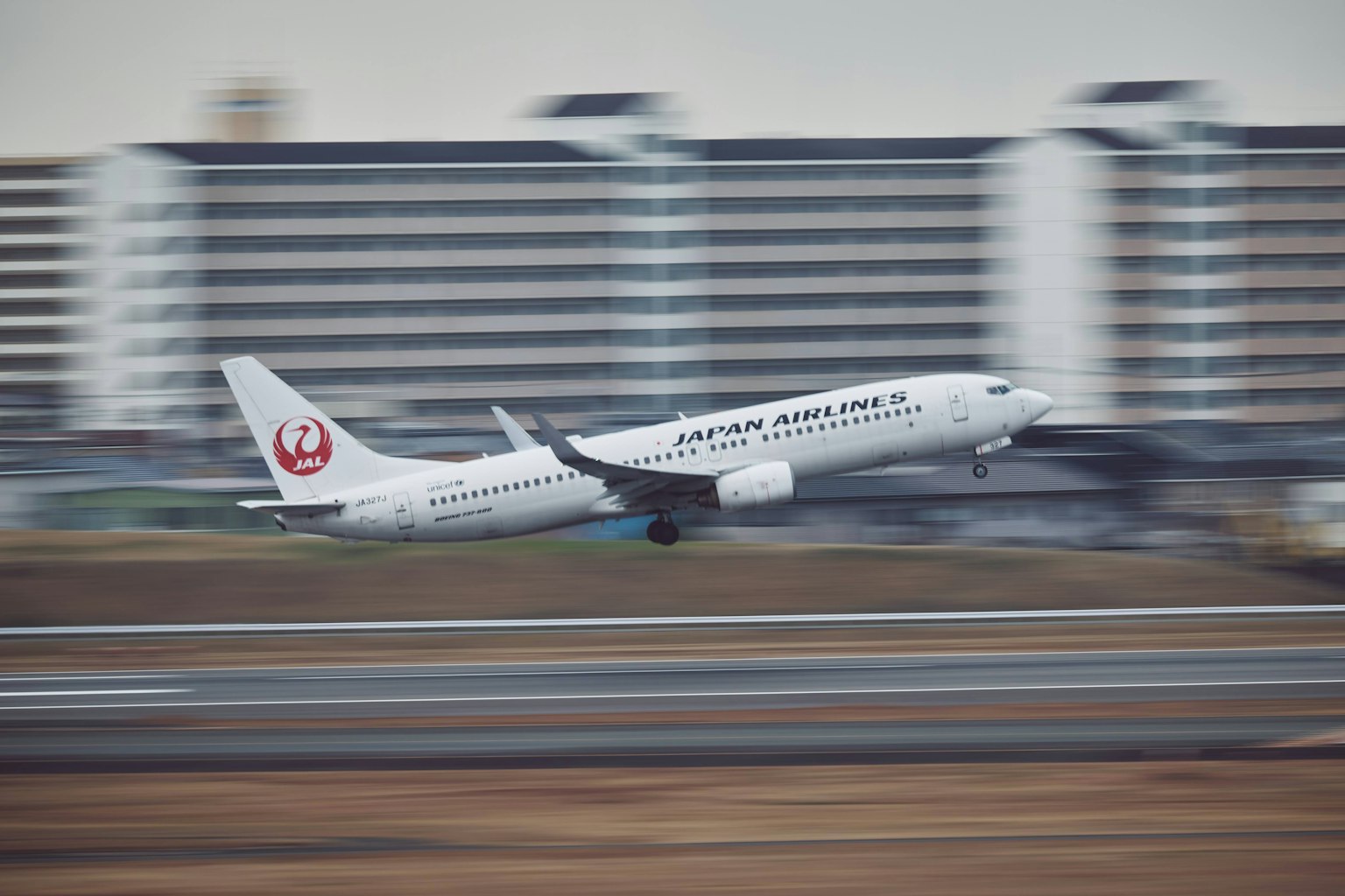 เครื่องบินสีขาวบินขึ้นจากรันเวย์มีตึกสูงอยู่เบื้องหลัง