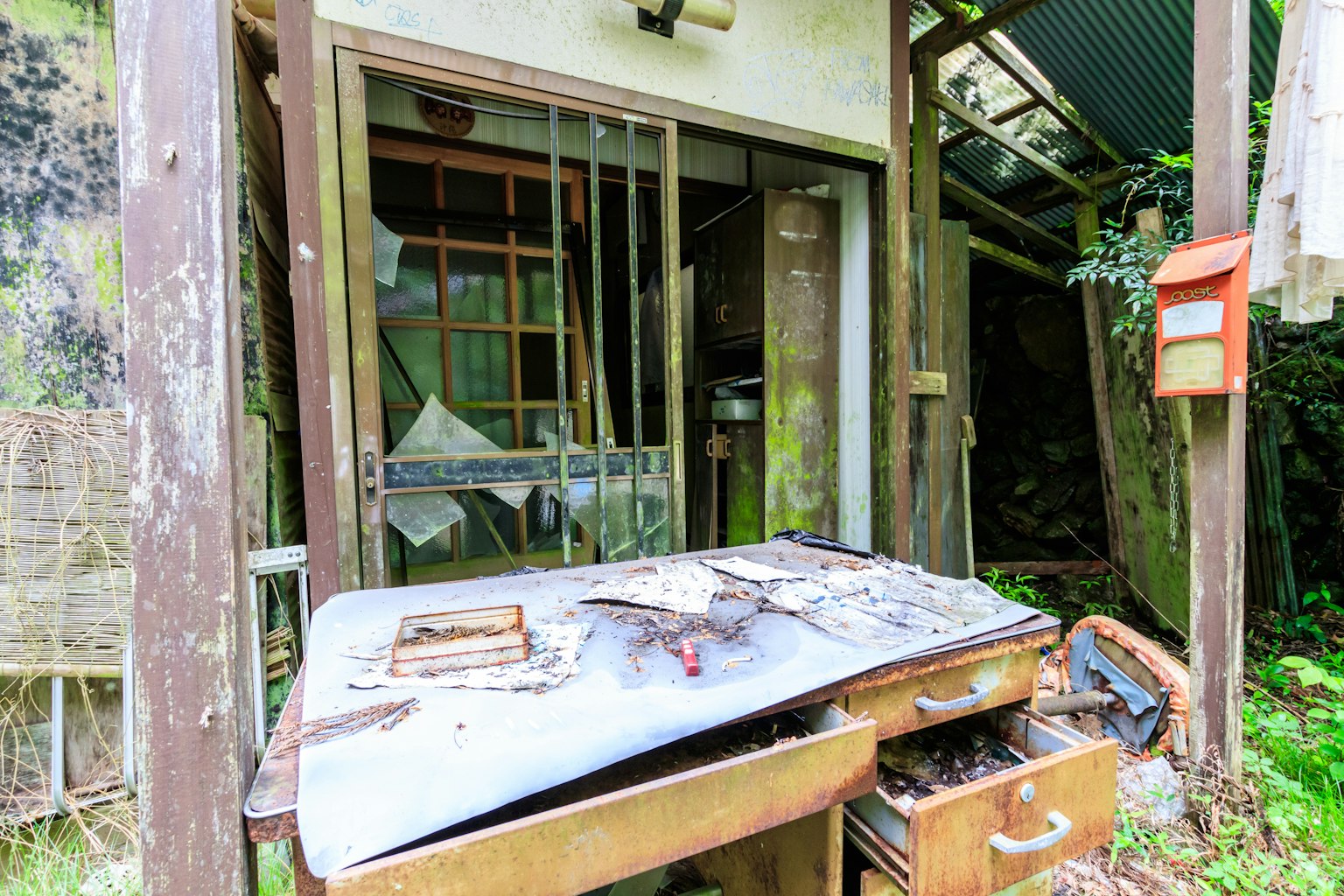 Eksterior rumah terbengkalai dengan meja usang dan vegetasi liar