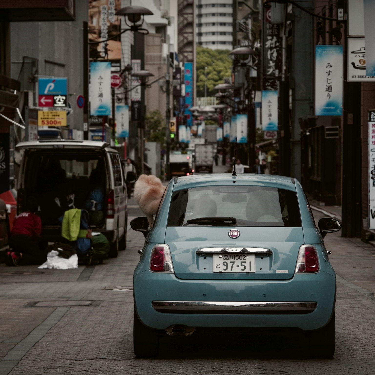 A blue car on a narrow street lined with commercial shops