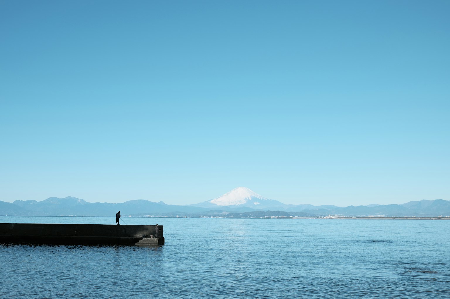 Una persona in piedi su un molo con cielo blu chiaro e oceano Monte Fuji visibile in lontananza