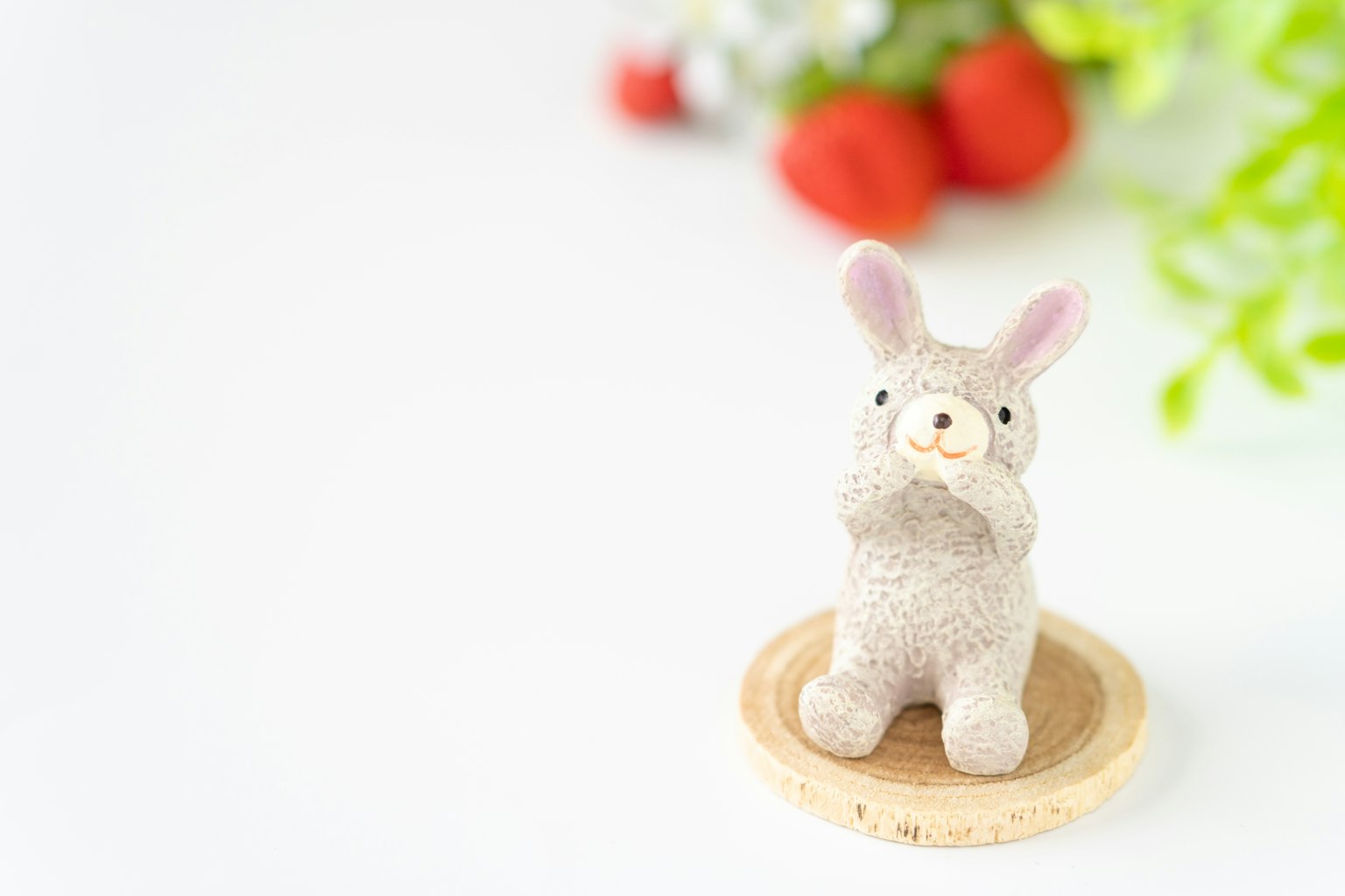 A small rabbit figurine sitting on a wooden plate with green plants and red fruits in the background