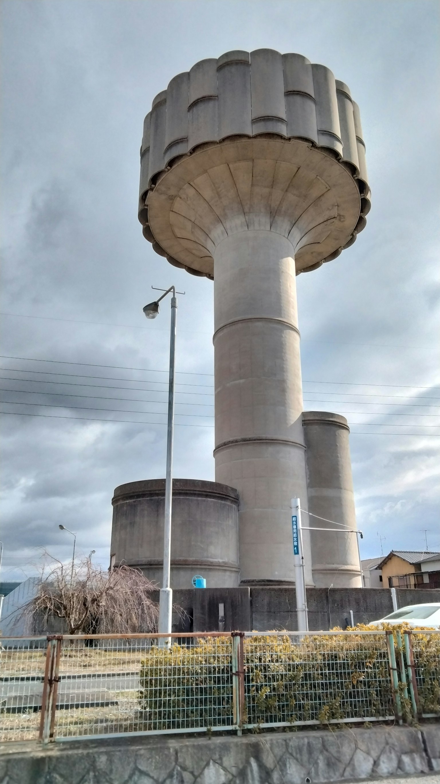 Una torre de agua alta con base cilíndrica y tapa grande rodeada de estructuras más pequeñas