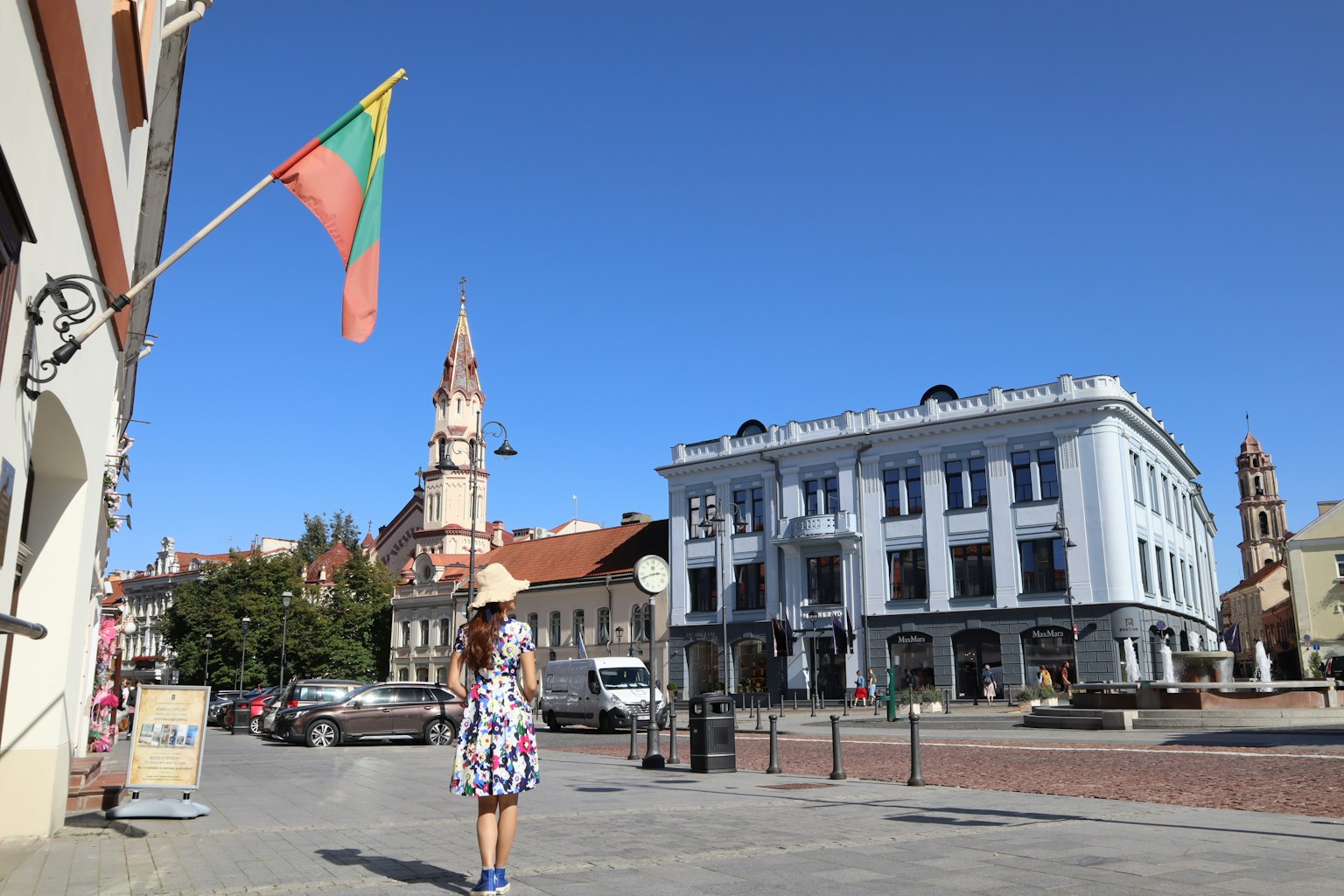 Une femme en robe à motifs marchant dans une place avec un drapeau lituanien et un bâtiment bleu
