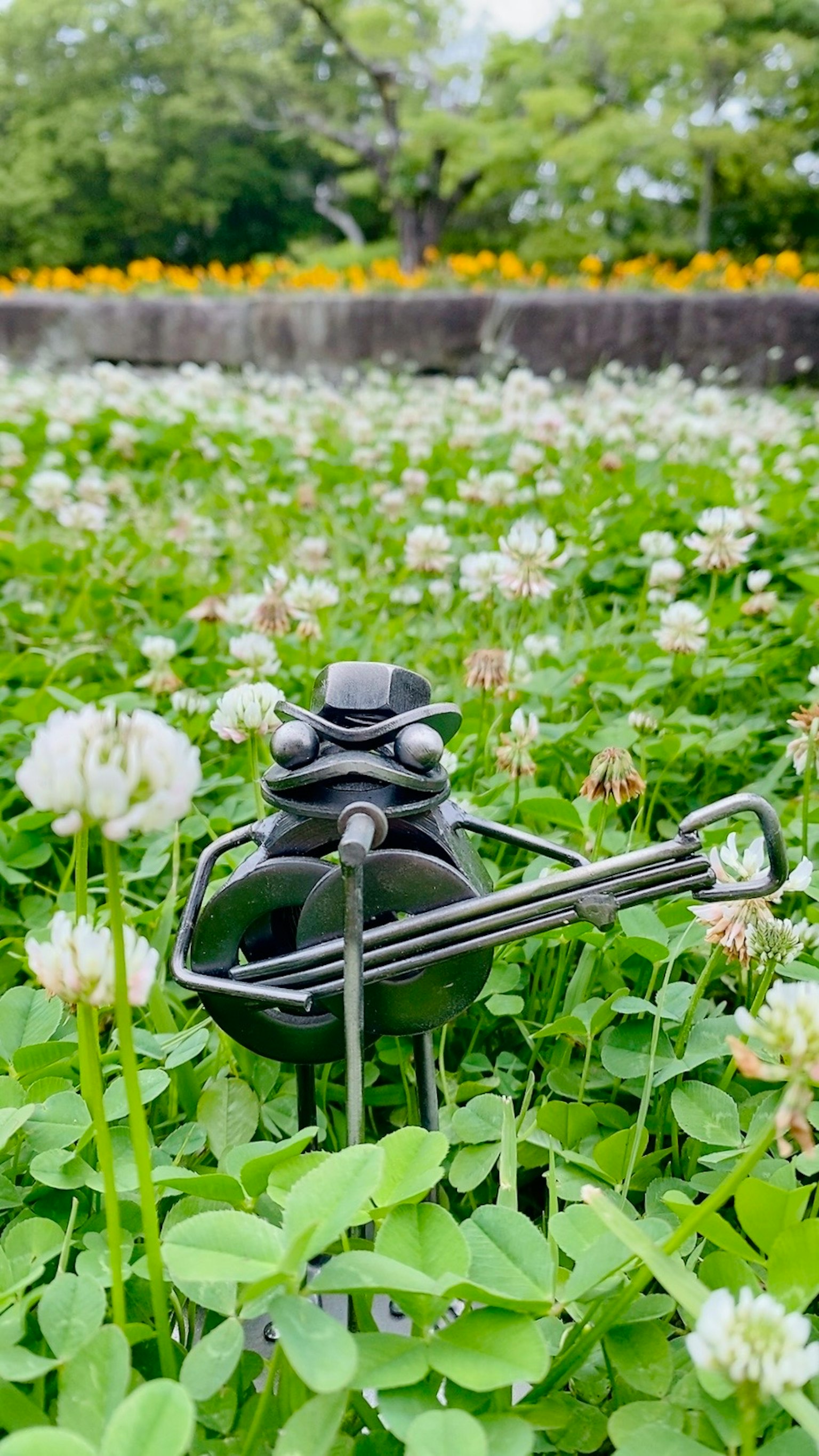Scultura di rana in metallo che si erge in un campo di erba verde e fiori di trifoglio bianco
