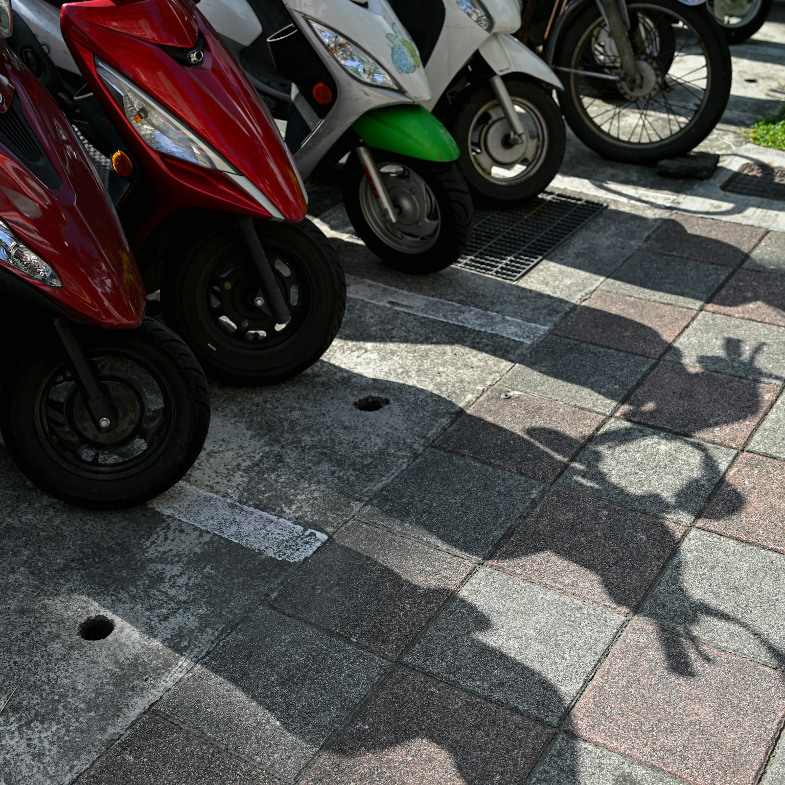 Zone de stationnement avec des scooters rouges et blancs projetant des ombres sur le pavé