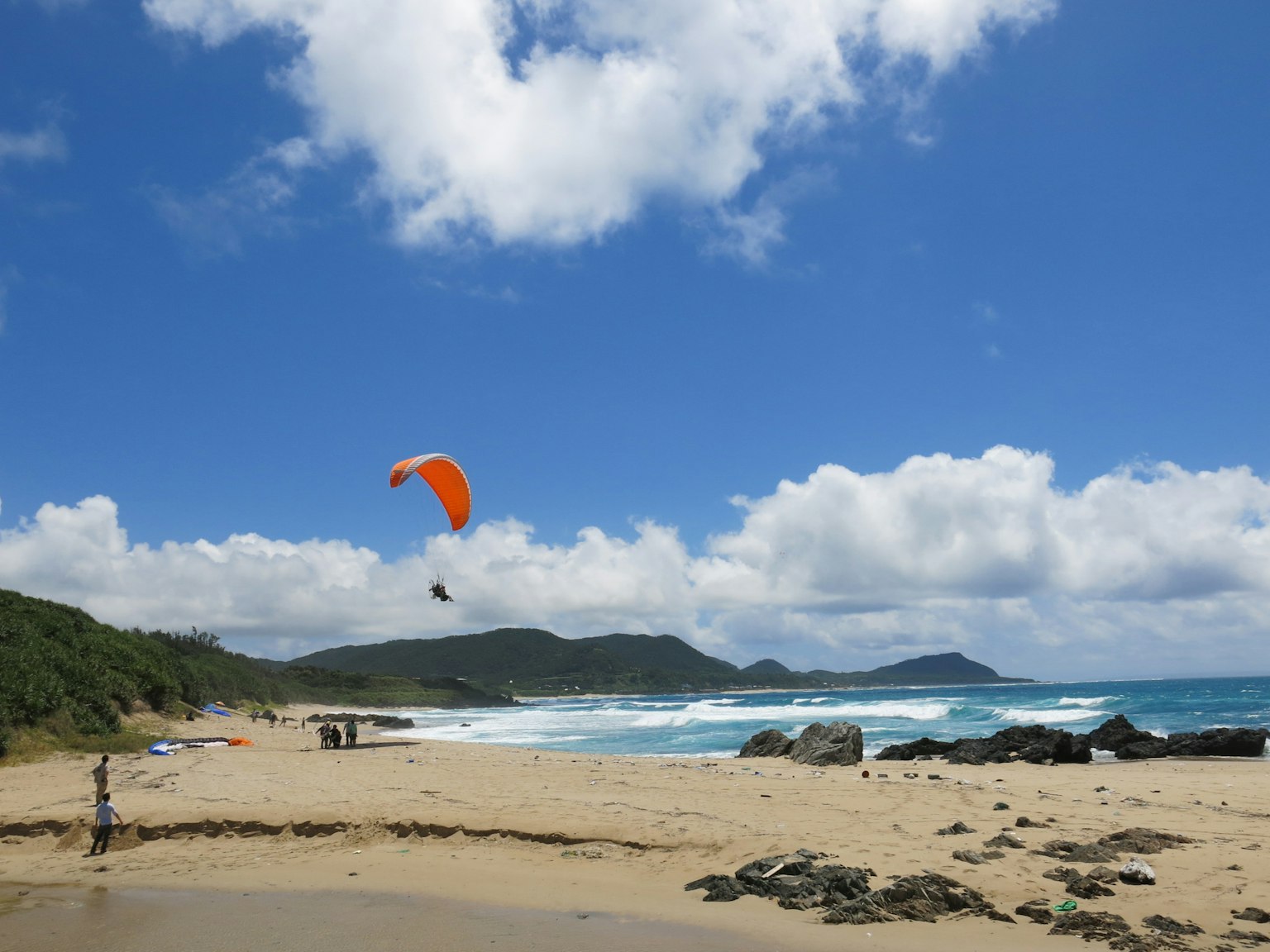 Pemandangan pantai dengan paralayang oranye terbang di bawah langit biru dan awan putih