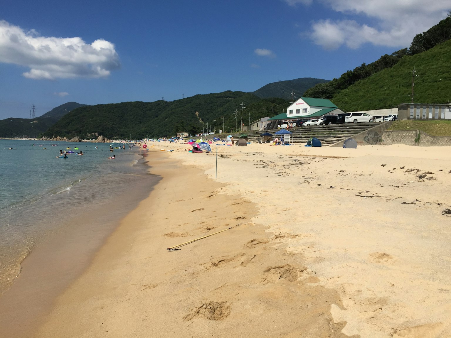 Vista panoramica della spiaggia con acqua blu e riva sabbiosa colline verdi e strutture sullo sfondo