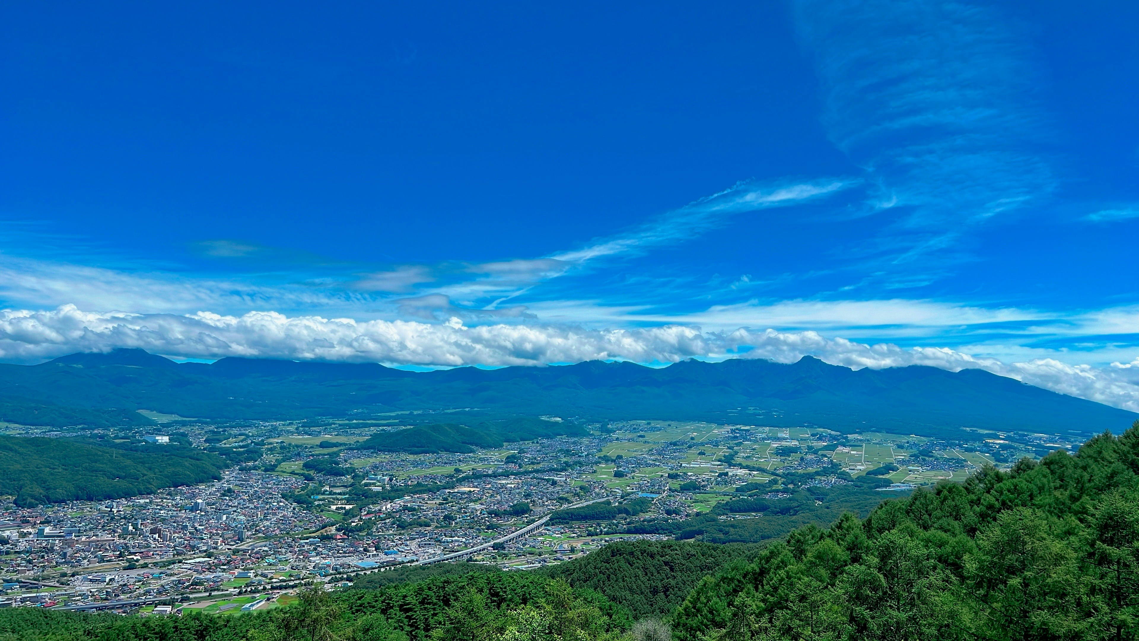 Vista de una ciudad desde colinas verdes bajo un cielo azul con montañas