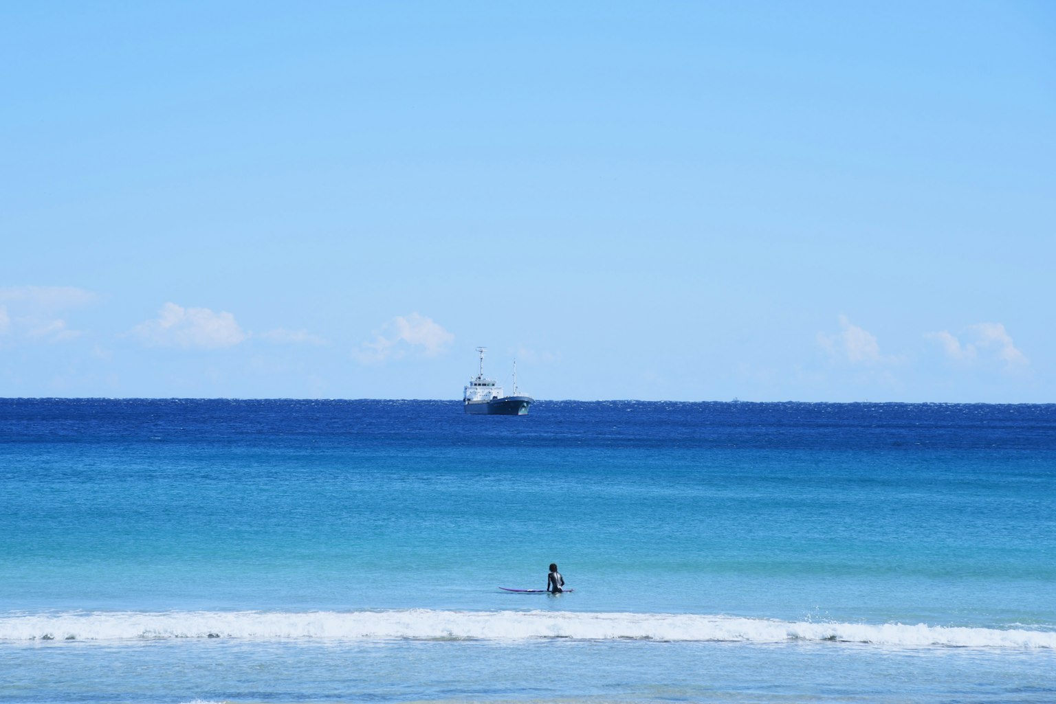 A person standing at the shoreline under a blue sky with a distant boat on the horizon