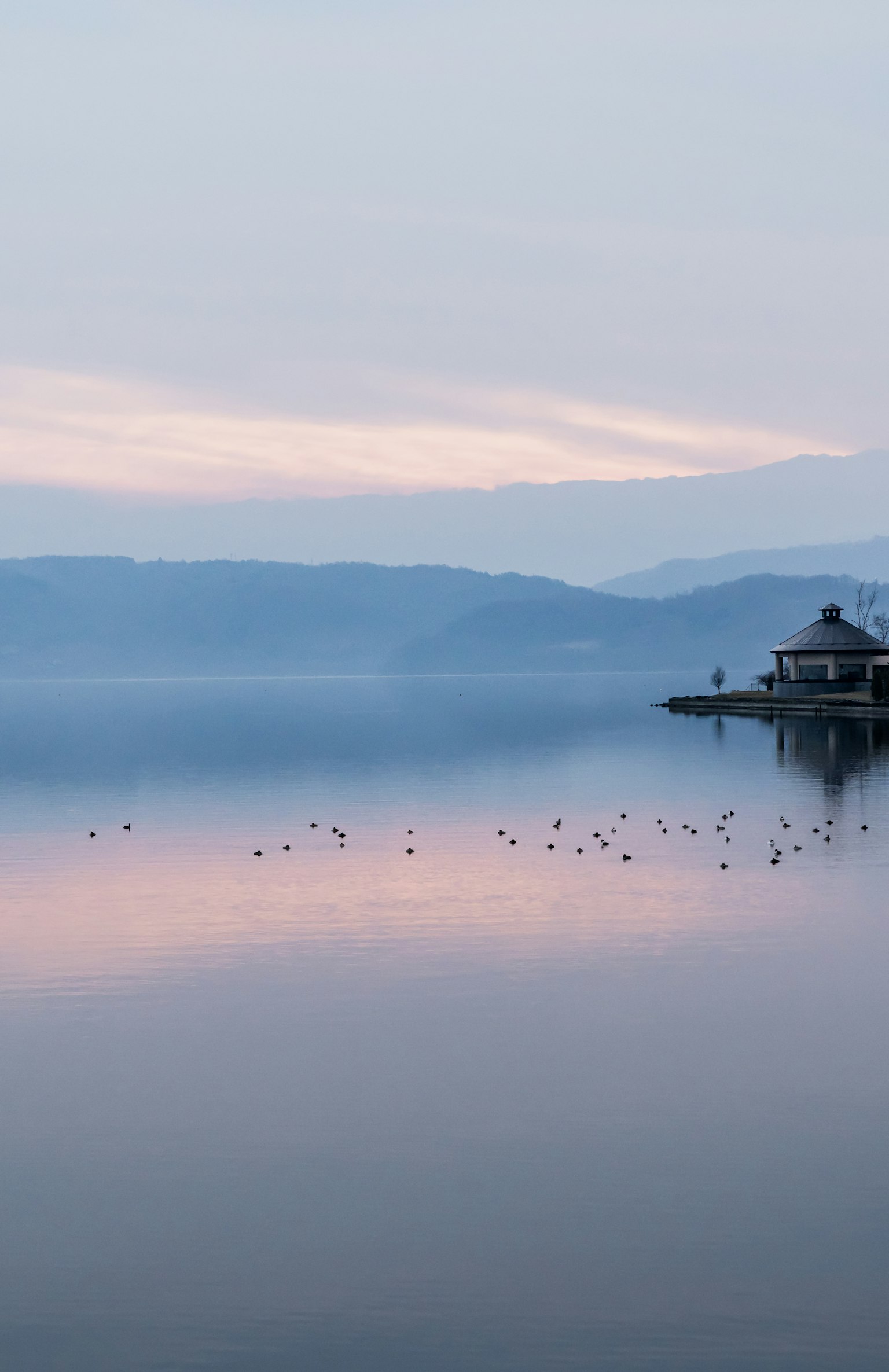 Ruhiger Blick auf den See mit einem kleinen Gebäude und sanften Farben des Himmels, die sich im Wasser spiegeln
