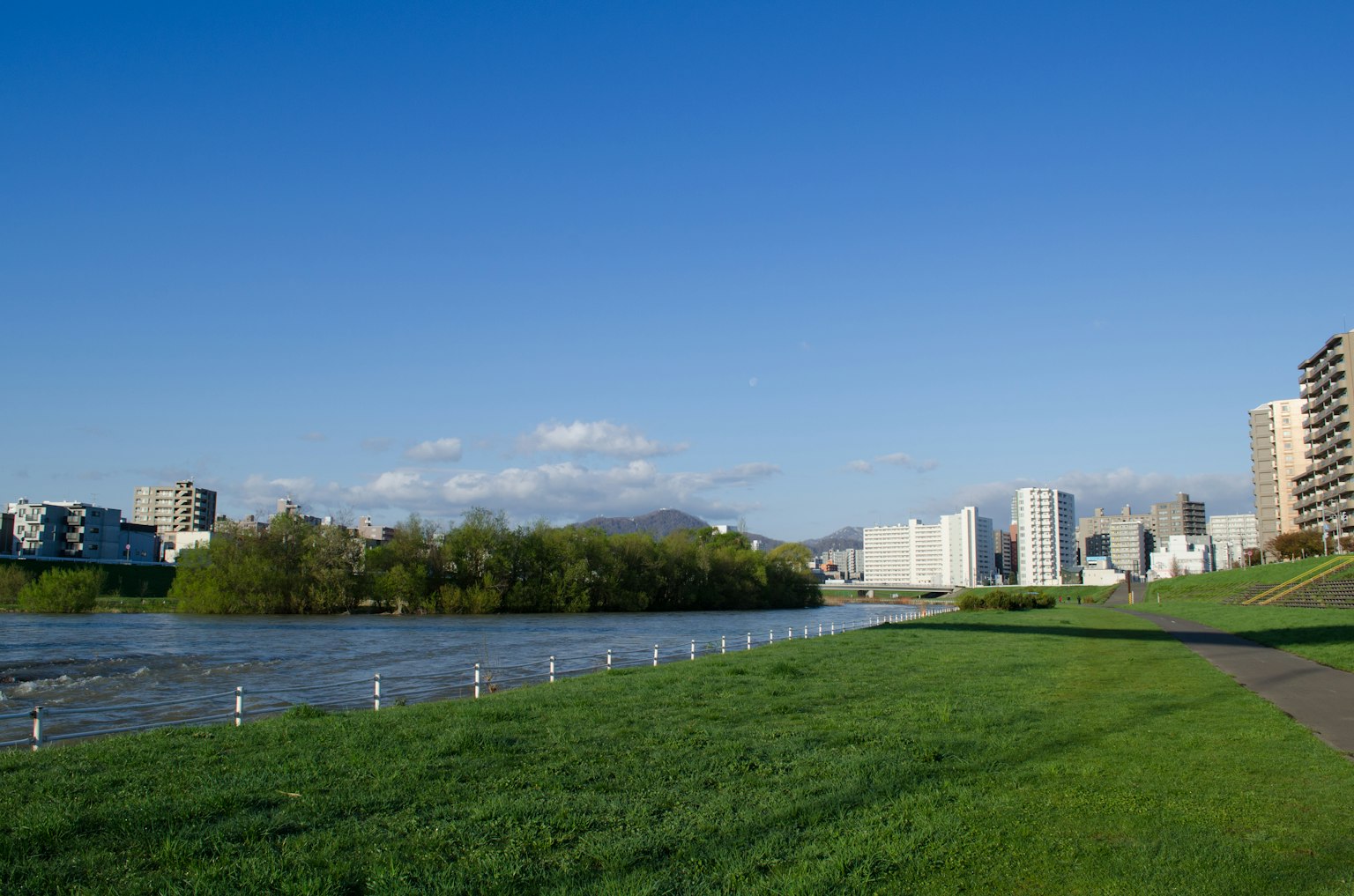 Scenic view of a river with buildings under a blue sky green grass and walkway