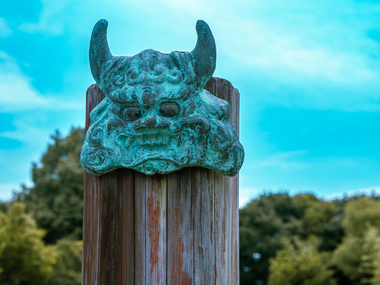 Bronzene Dämonenkopfskulptur auf einem Holzpfosten vor natürlichem Hintergrund