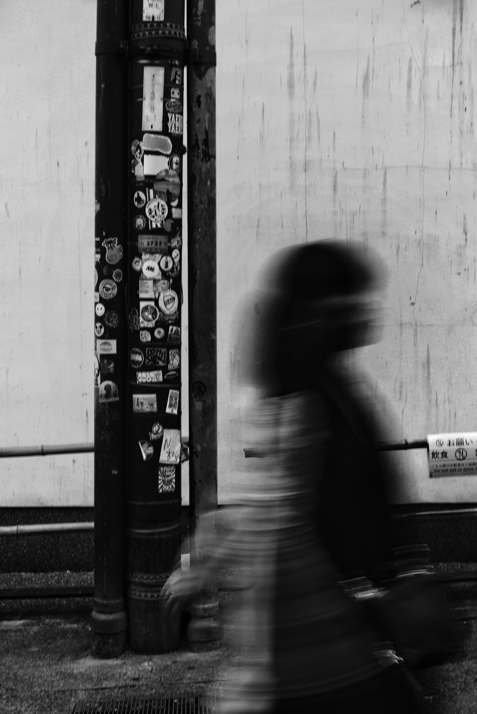 Black and white urban scene featuring a moving person and a pole covered in stickers