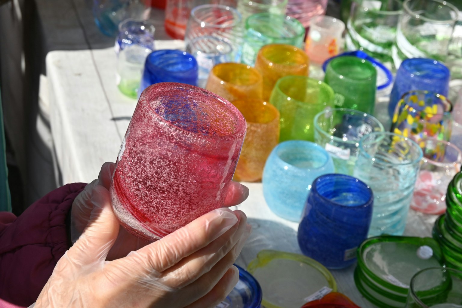 Gros plan sur une main tenant une tasse en verre colorée parmi divers objets en verre