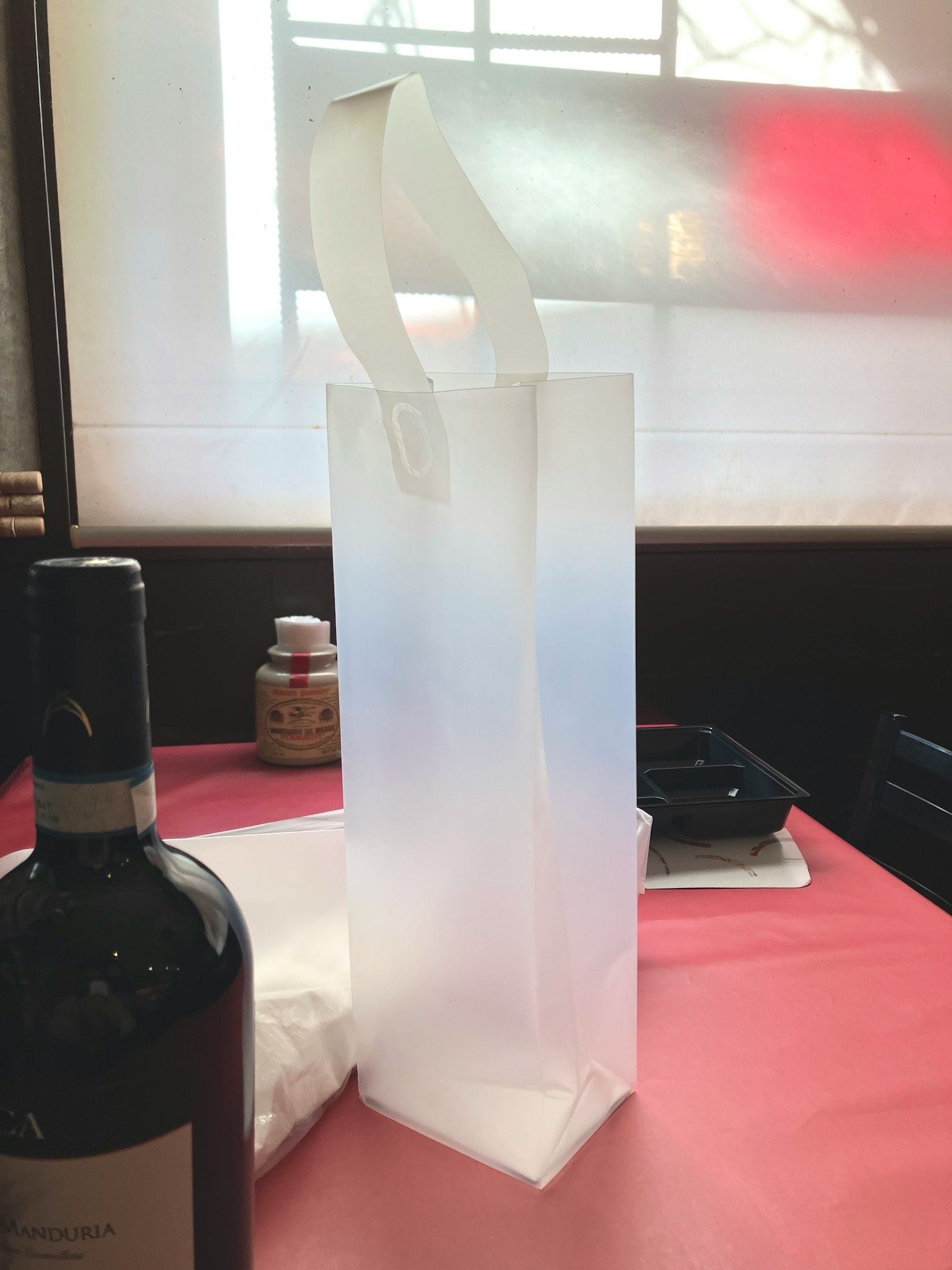 Una borsa di carta bianca è posata su un tavolo con una tovaglia rossa e una bottiglia di vino sullo sfondo