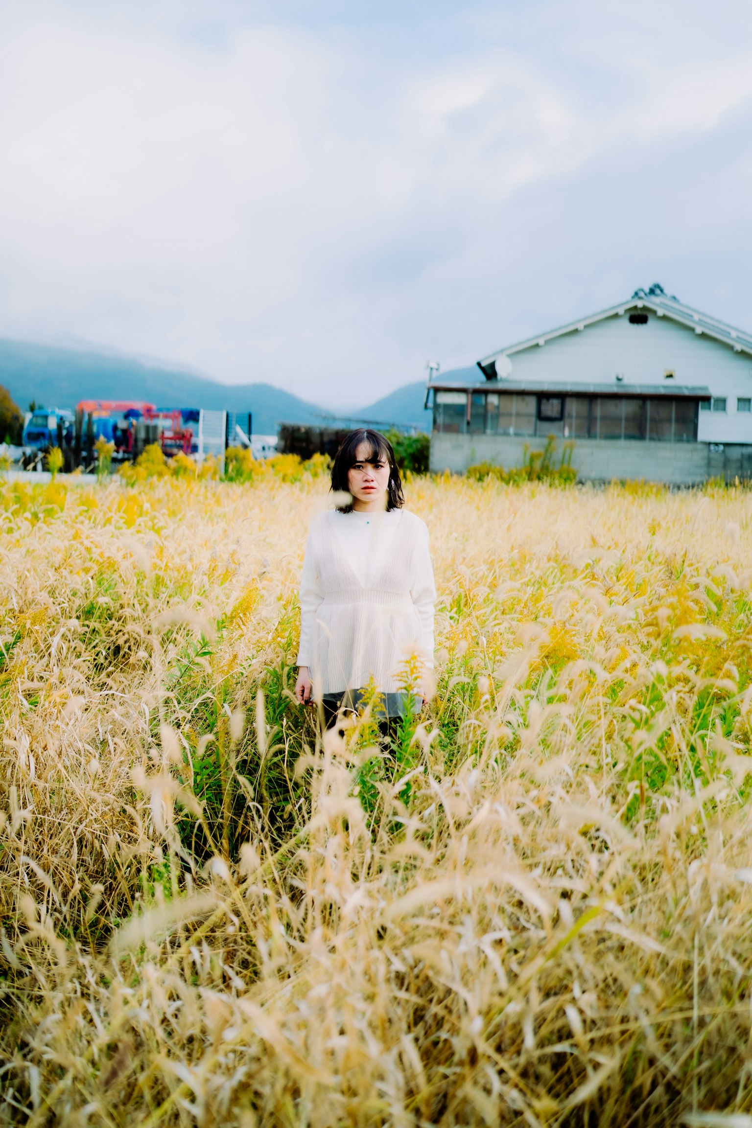 女人站在寬闊的田野中，背景是房子和山脈