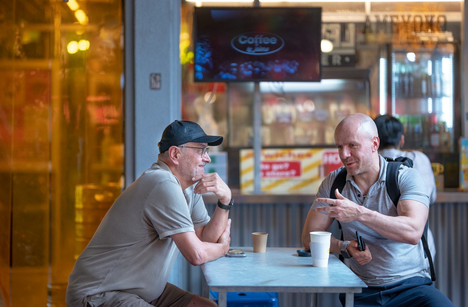 Zwei Männer, die in einem Café mit Kaffeetassen auf dem Tisch plaudern helles Licht und ein Essensstand im Hintergrund