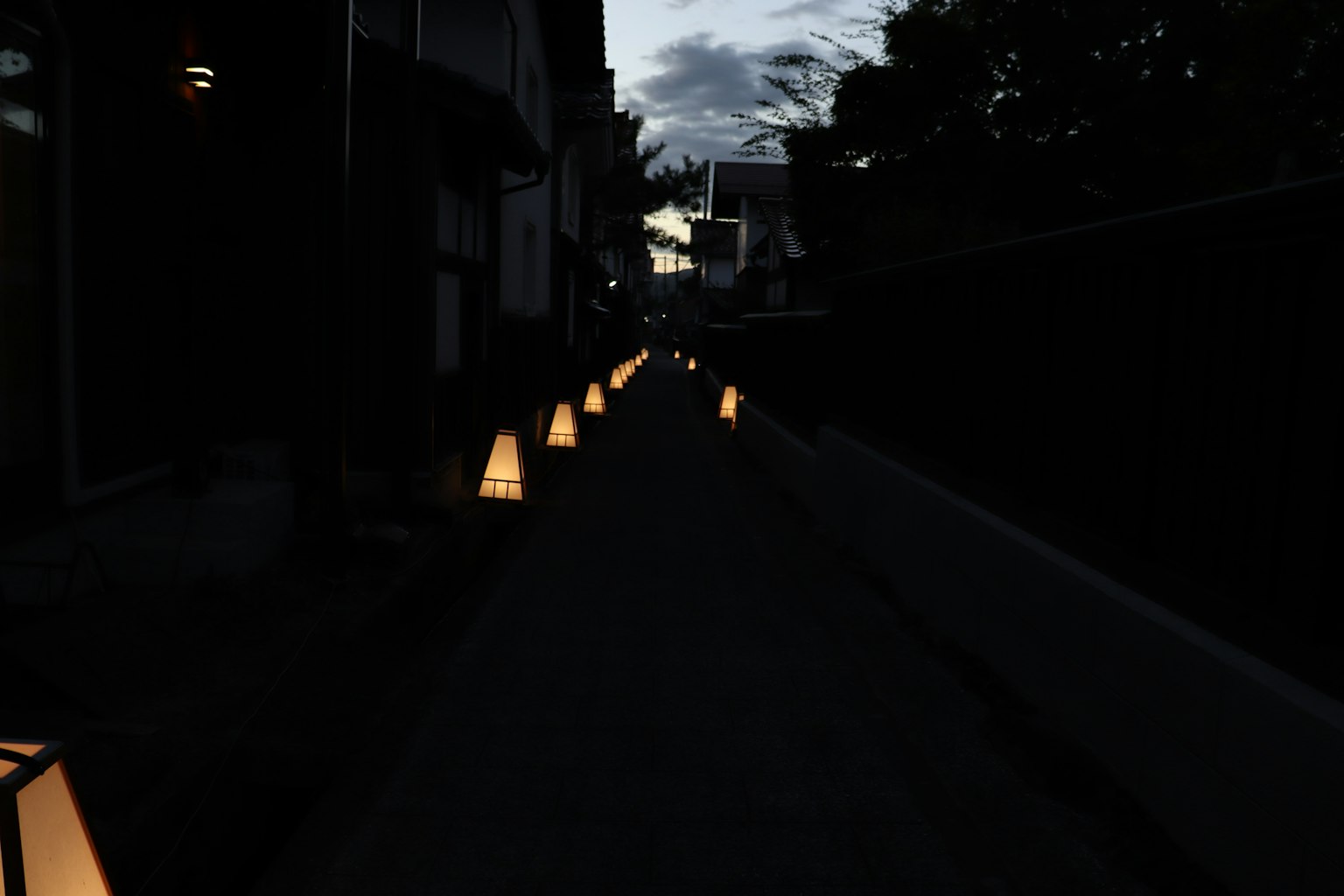 ทางเดินที่มีแสงสลัวเรียงรายไปด้วยโคมไฟที่ส่องสว่างและอาคารรอบข้าง