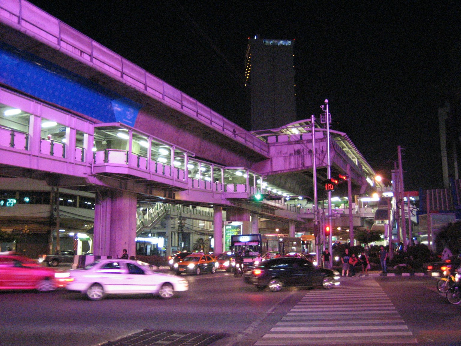 Escena urbana nocturna con tren elevado y cruce concurrido iluminado de rosa