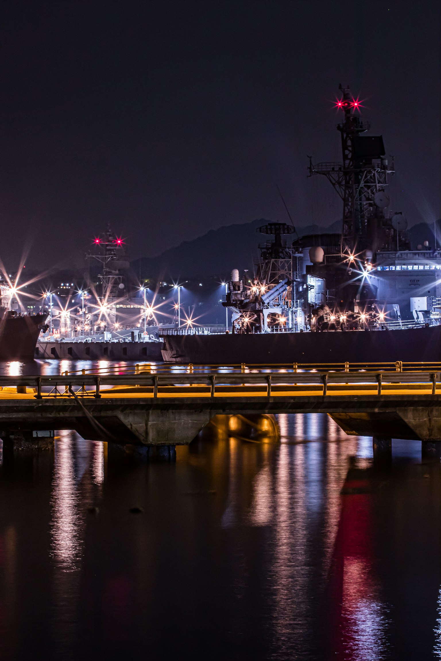 Vista nocturna de un puerto con barcos iluminados y reflejos del puente fondo industrial