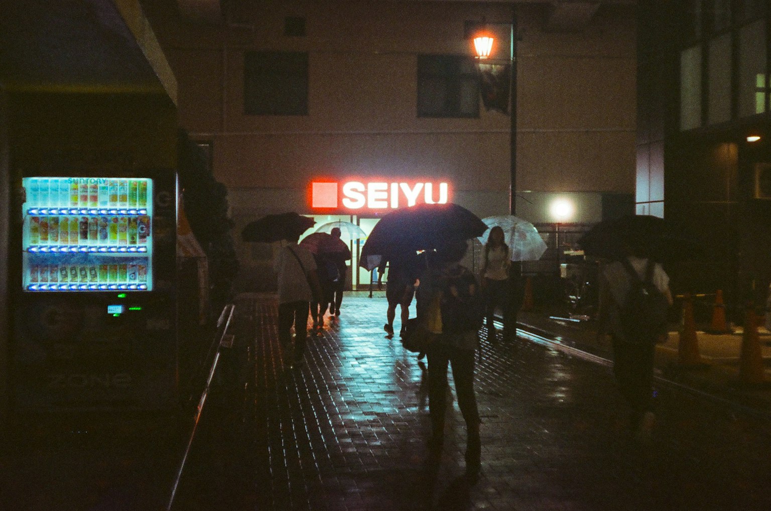 Menschen mit Regenschirmen, die im Regen gehen, beleuchtet vom hellen SEIYU-Schild