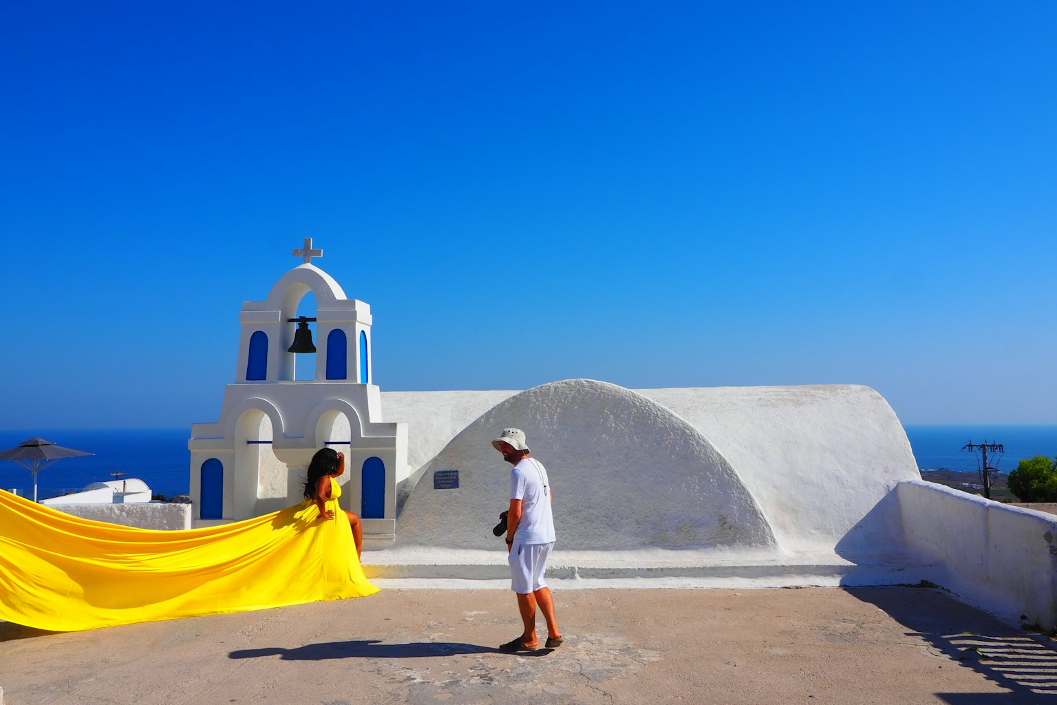 Une femme en robe jaune pose pour une photo avec un homme en blanc près d'une église blanche sous un ciel bleu