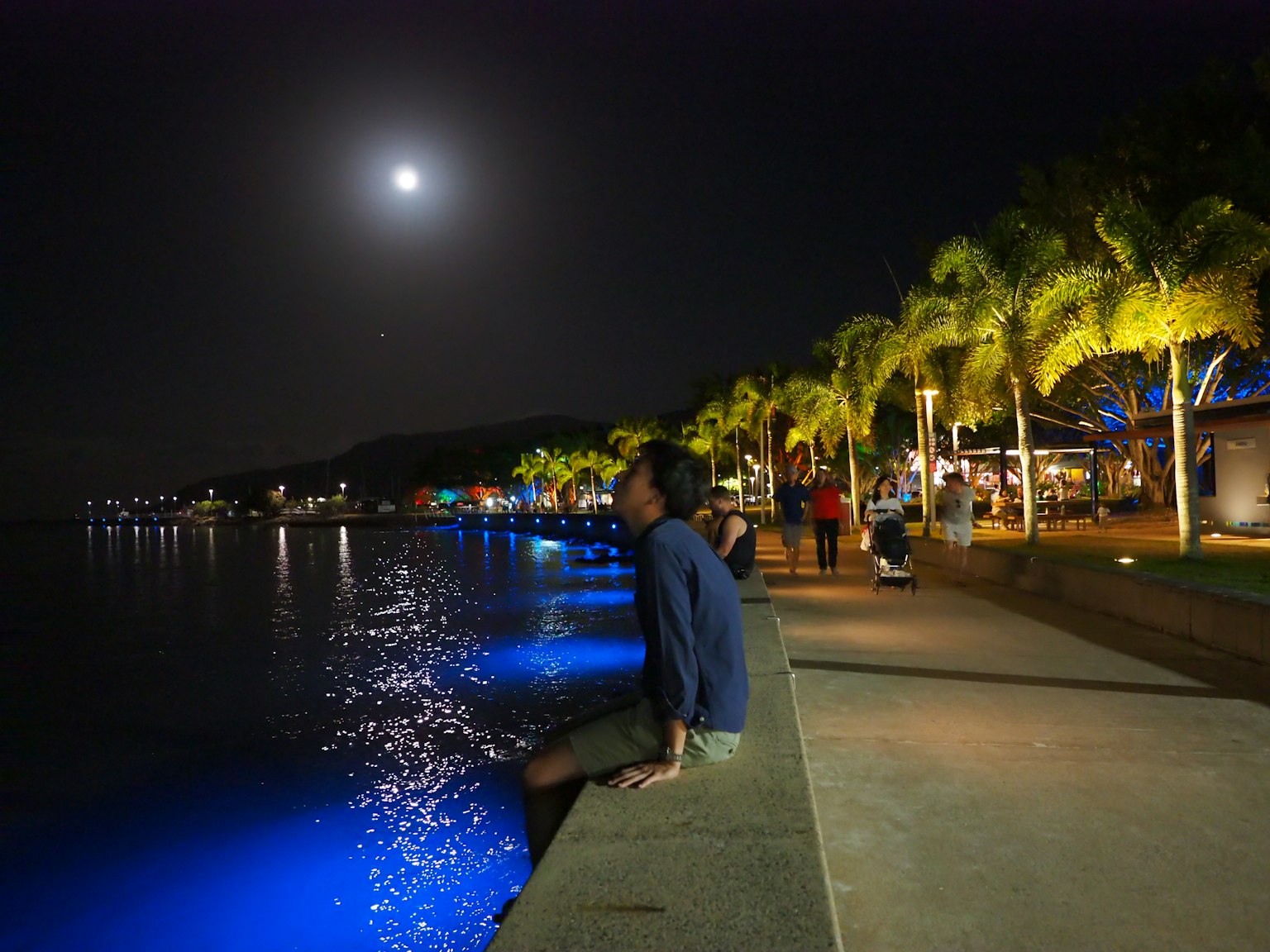 Un homme assis au bord de la mer la nuit sous la lumière de la lune avec de l'eau éclairée en bleu