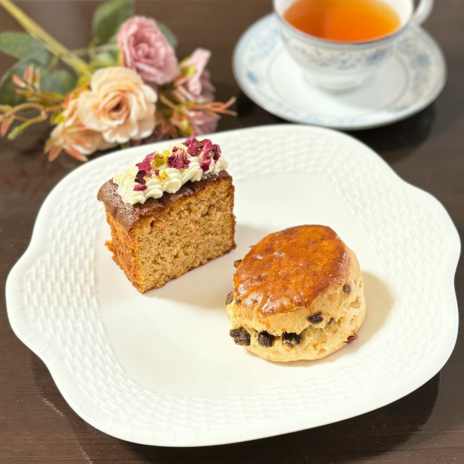 Un piatto con una fetta di torta guarnita di panna e uno scone con uvetta accanto a una tazza di tè