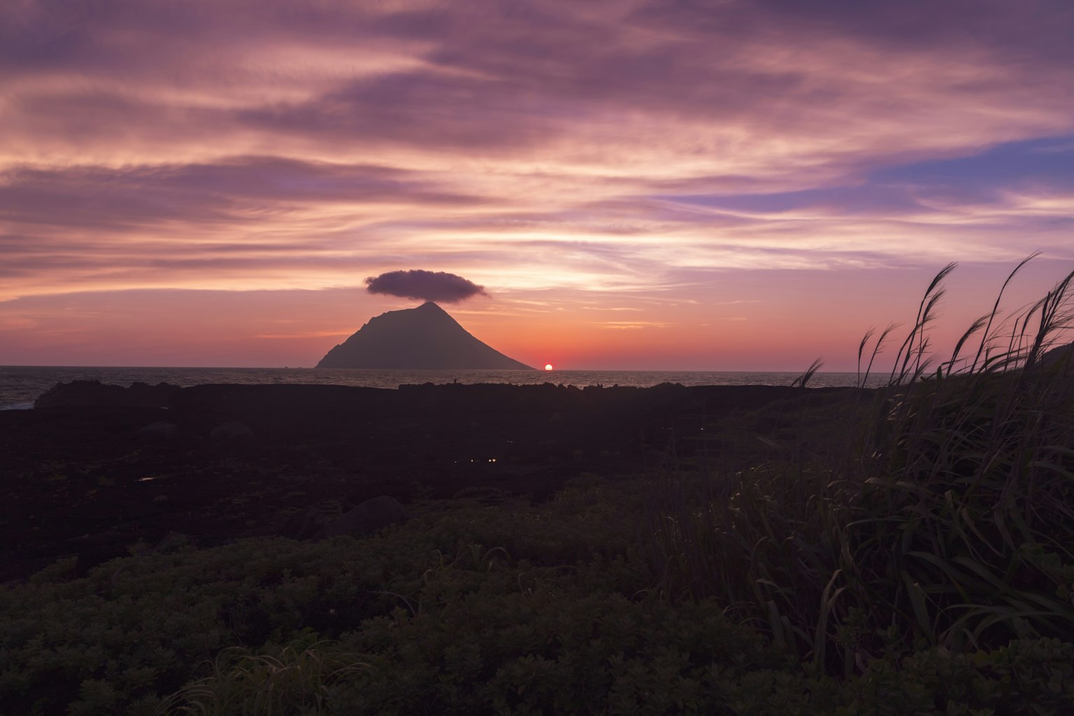 Silhouette of a mountain with a cloud on top against a sunset sky