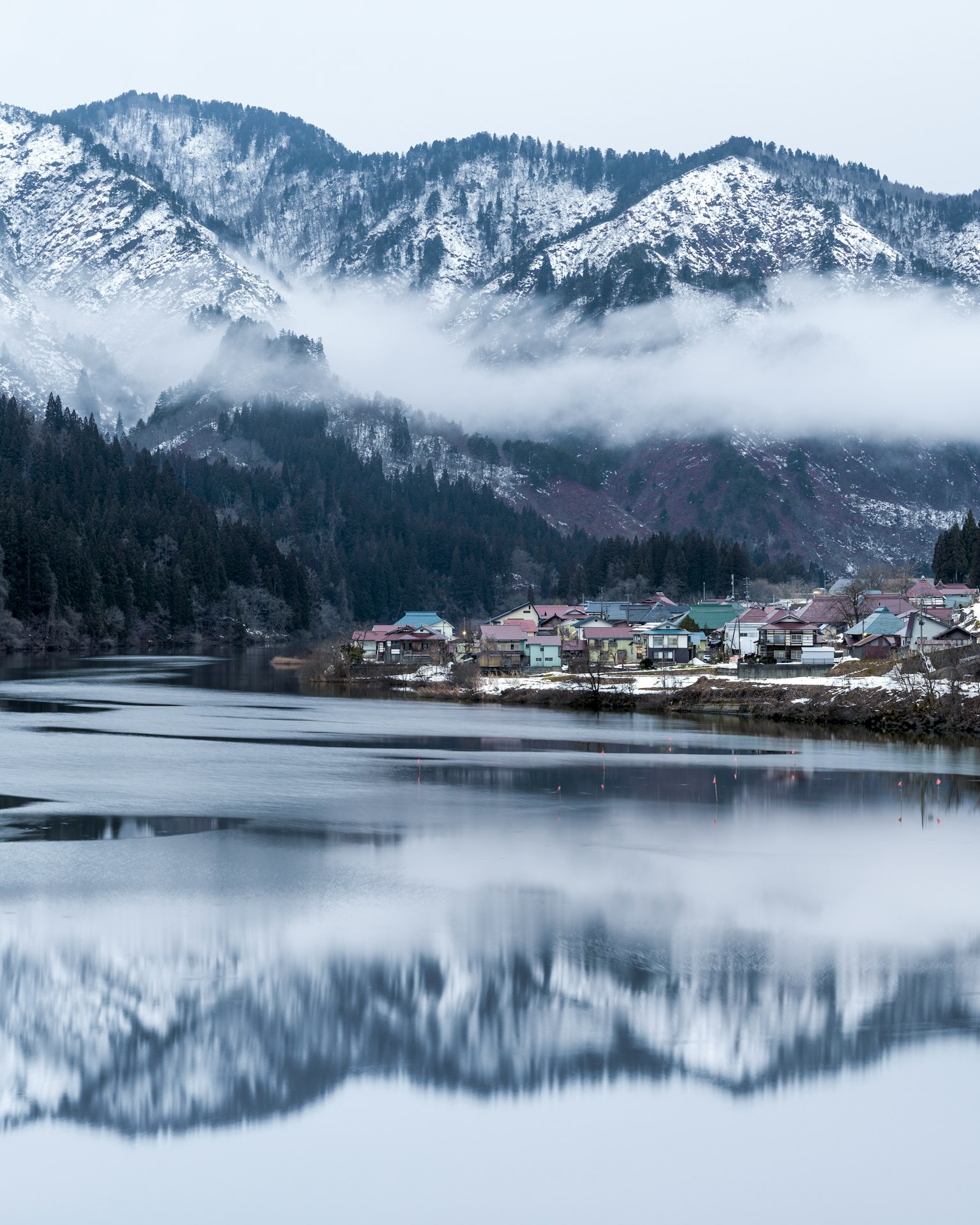 被雪覆盖的山脉和宁静的湖泊景观反射村庄房屋和雾气