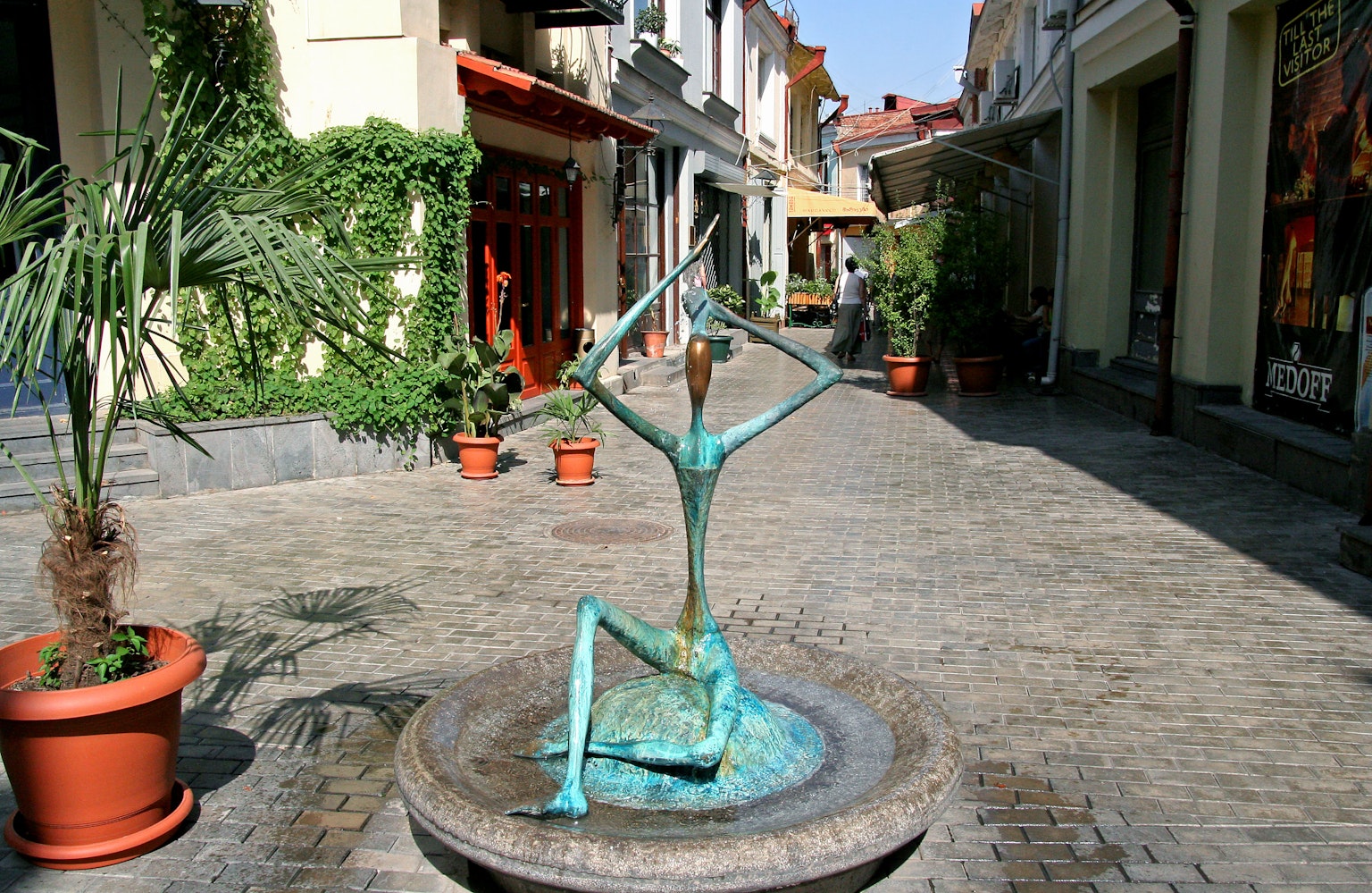 Escultura de bronce en una calle adoquinada con tiendas y plantas en maceta