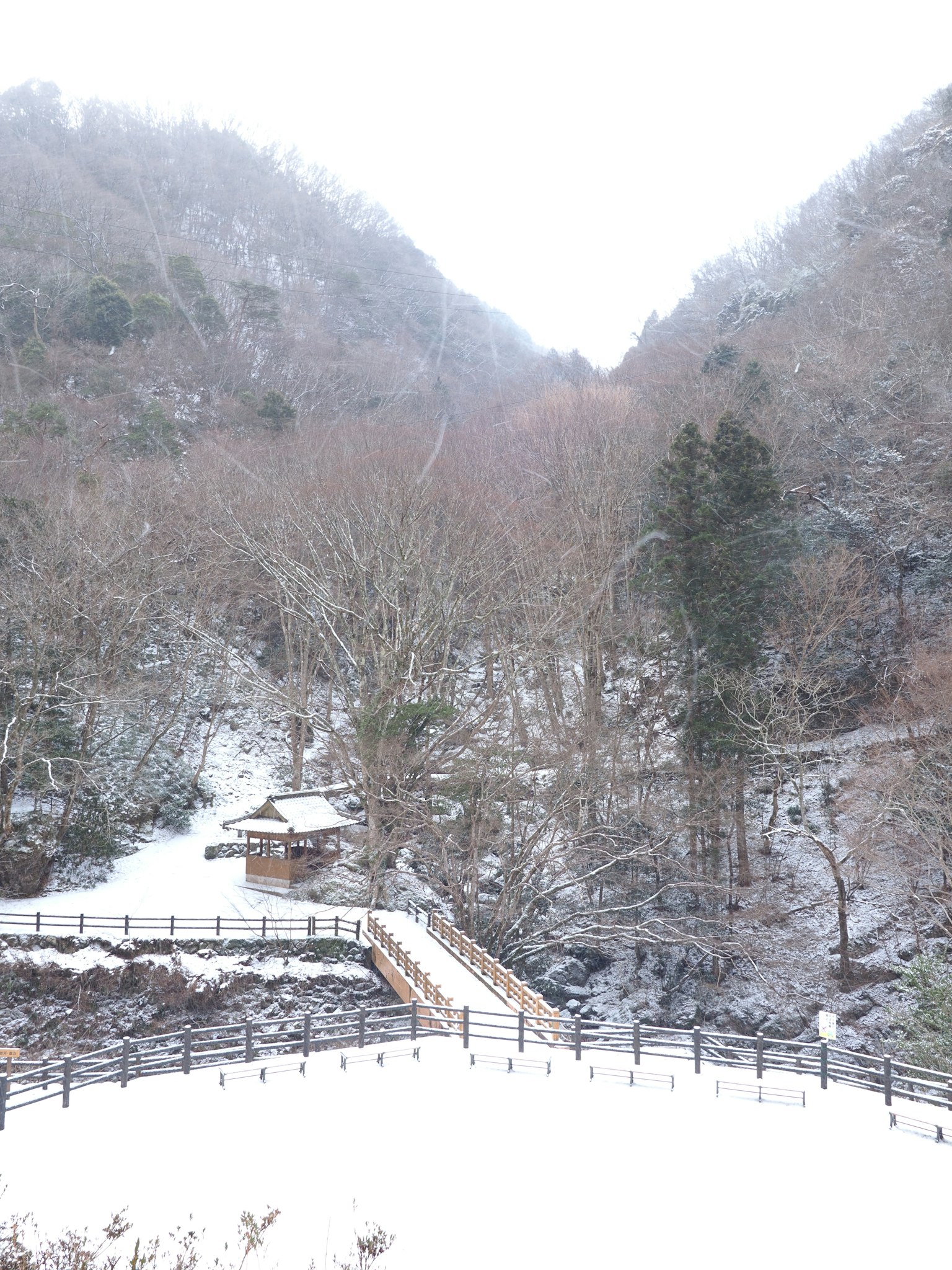 Escena de montañas cubiertas de nieve y río con una cabaña y un puente de madera