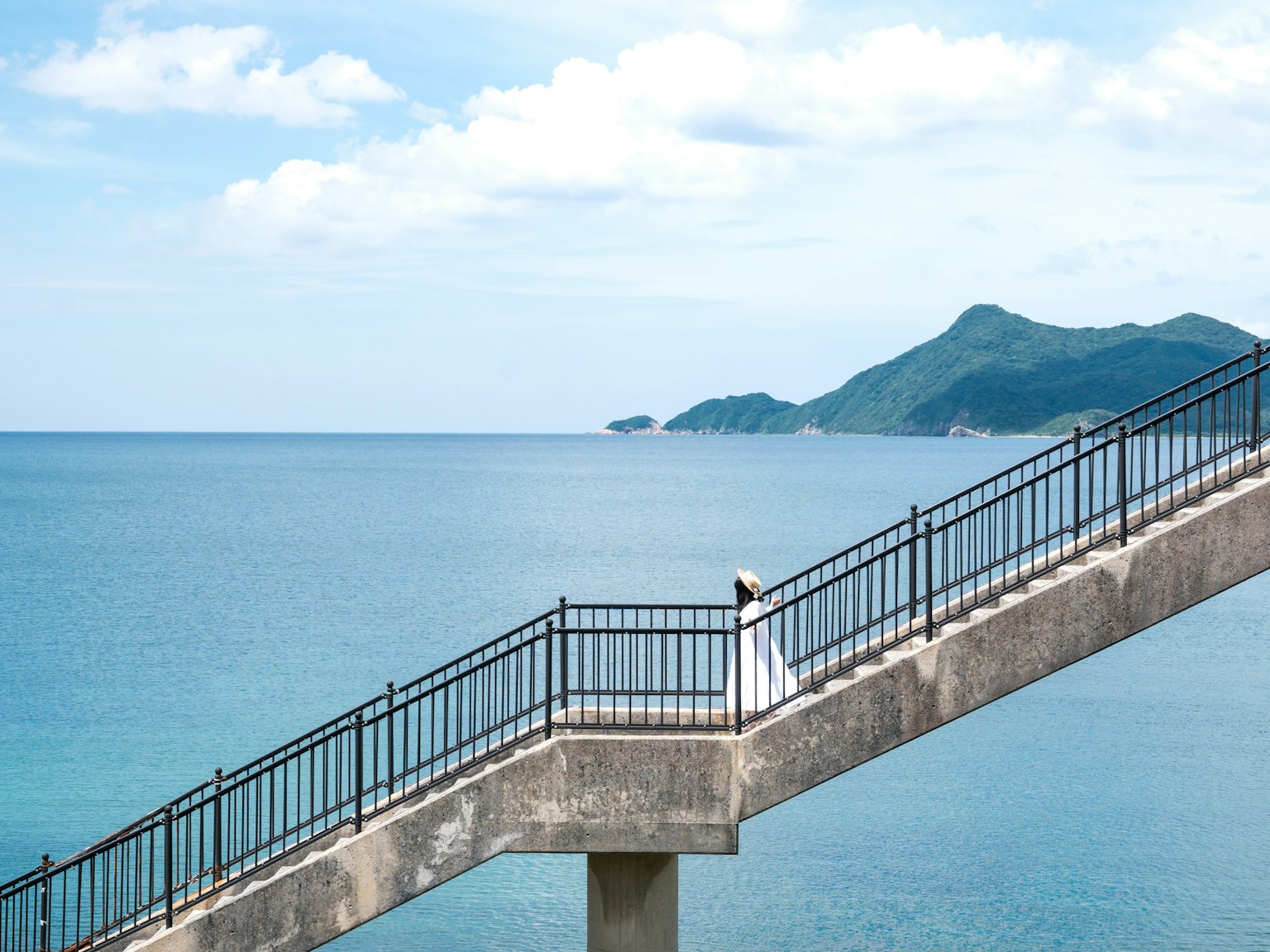 一位穿著白色裙子的女性在寧靜的藍色海洋旁的樓梯上走