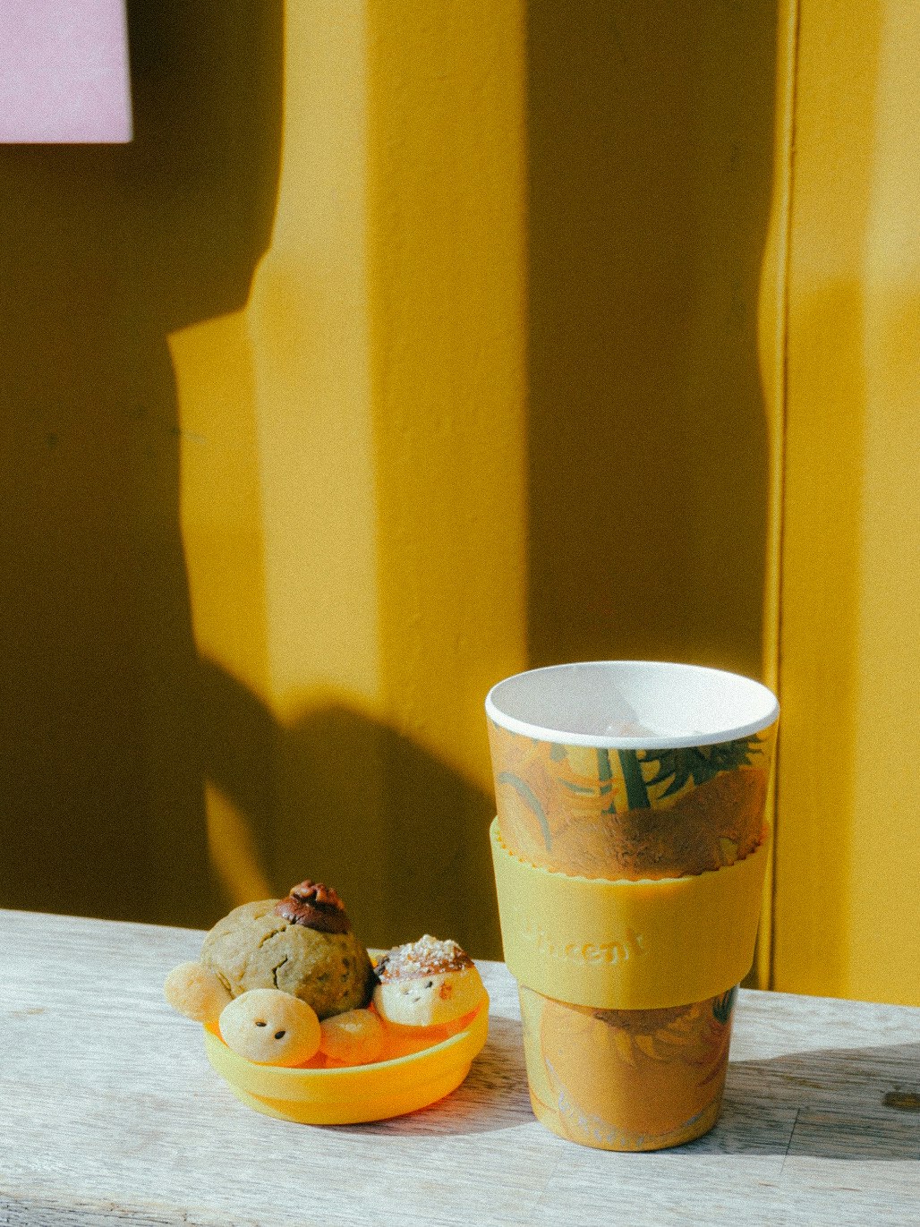 Coffee cup with a yellow sleeve beside a plate of cute snacks against a yellow background