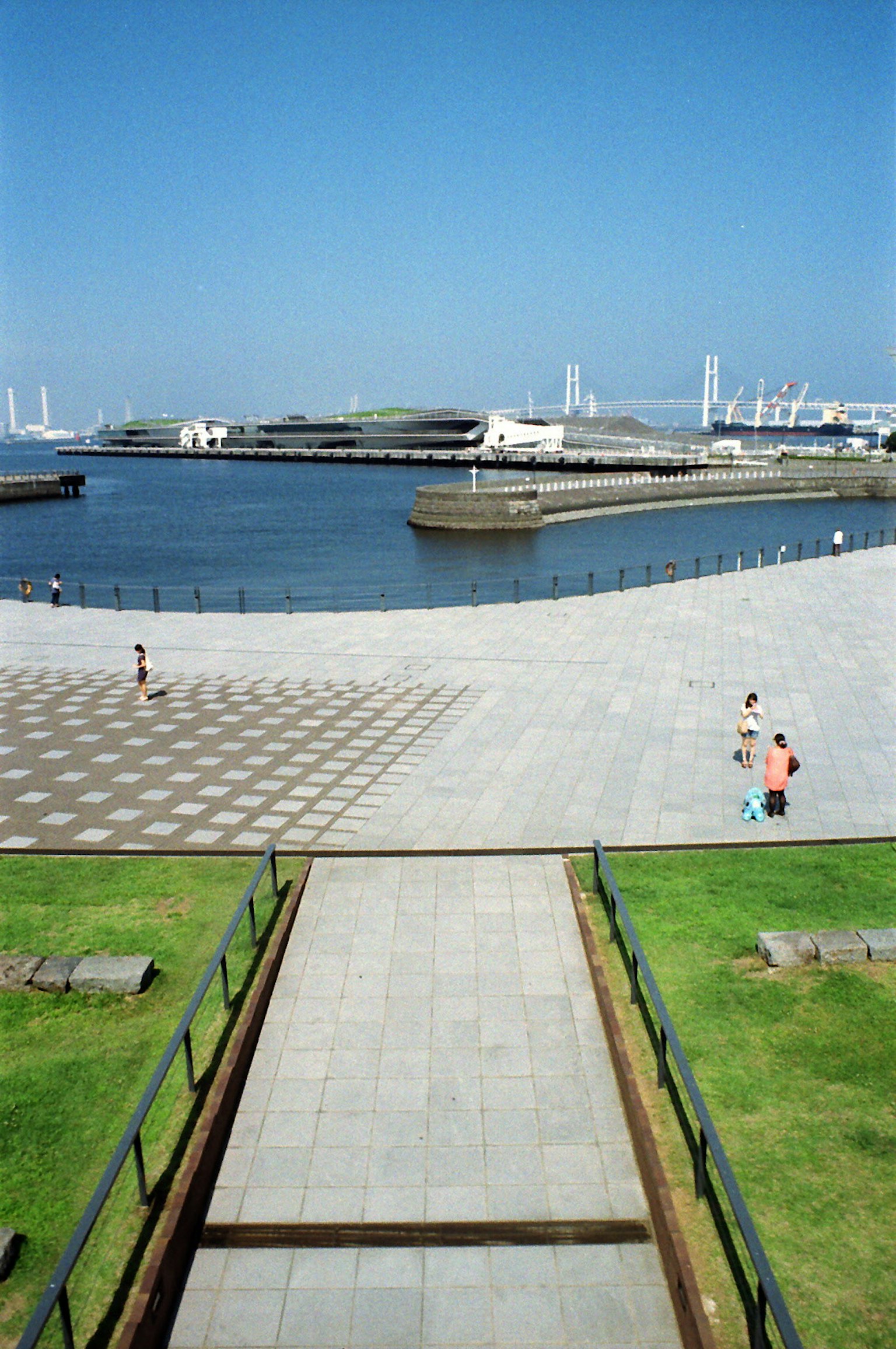 Vista escénica con cielo azul y agua que presenta una gran plaza y un camino con personas caminando