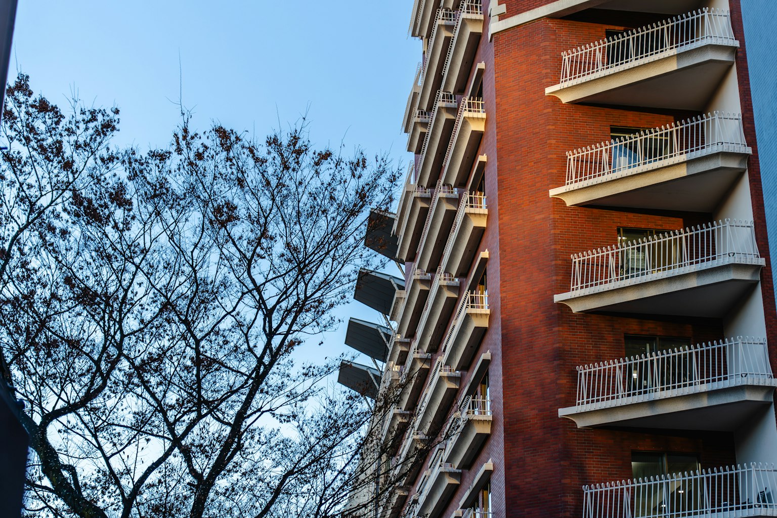 Vista lateral de un edificio de ladrillo rojo con balcones y la silueta de un árbol