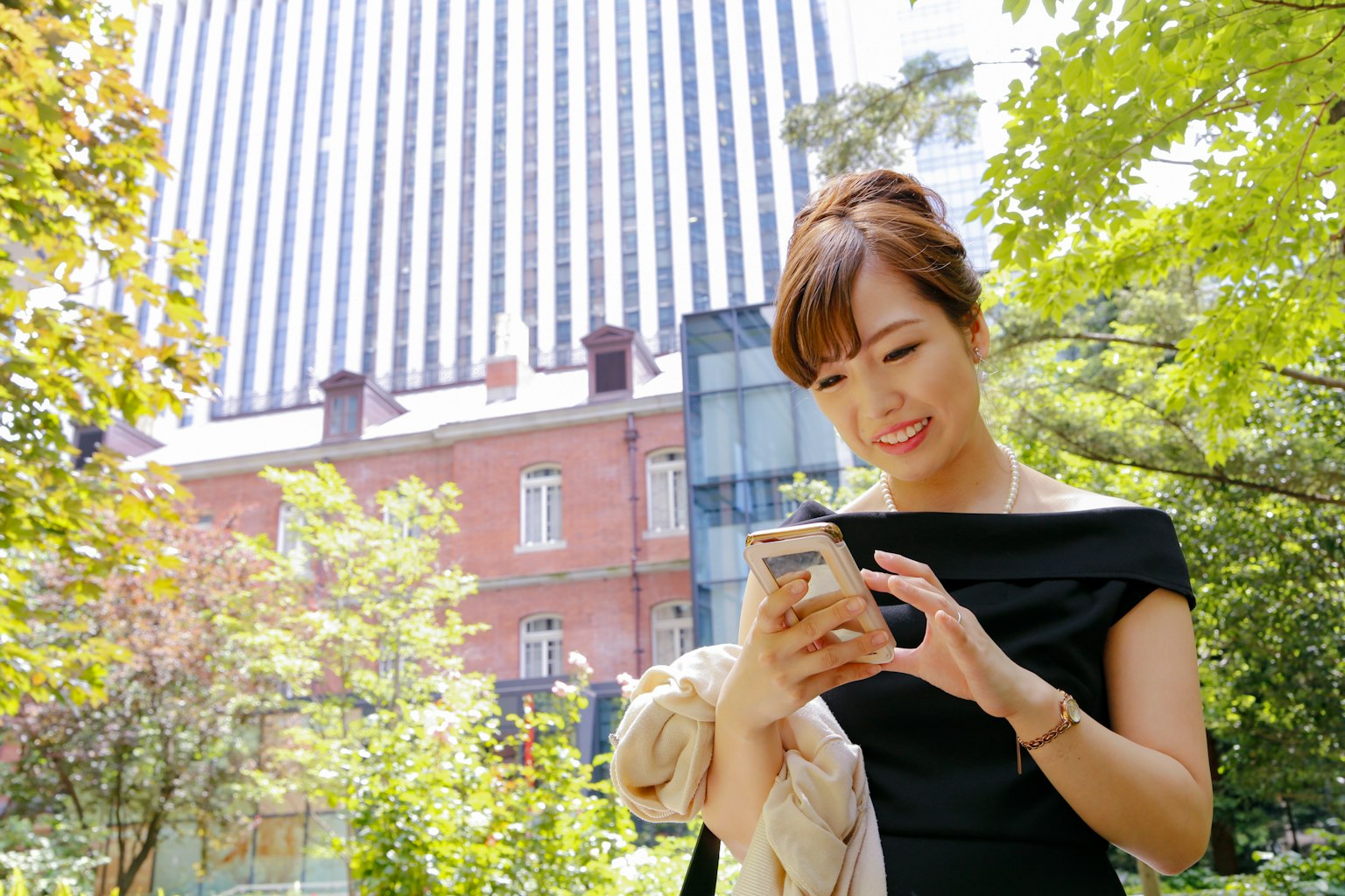 Woman using smartphone in a park surrounded by greenery and a nearby building