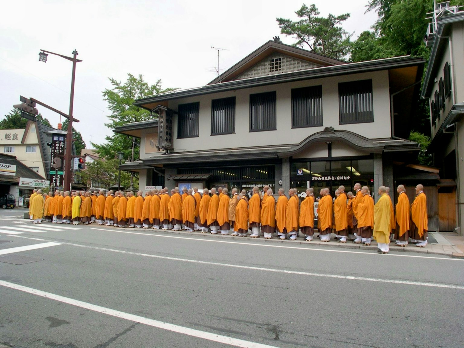 พระสงฆ์ในจีวรสีส้มเรียงแถวอยู่บนถนนมีอาคารแบบดั้งเดิมอยู่เบื้องหลัง