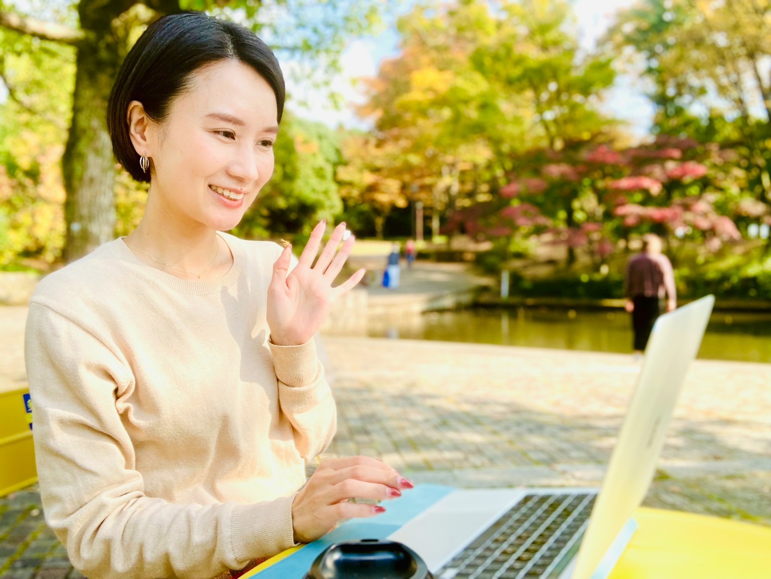 Junge Frau, die lächelt und winkt, während sie in einem Park einen Laptop benutzt