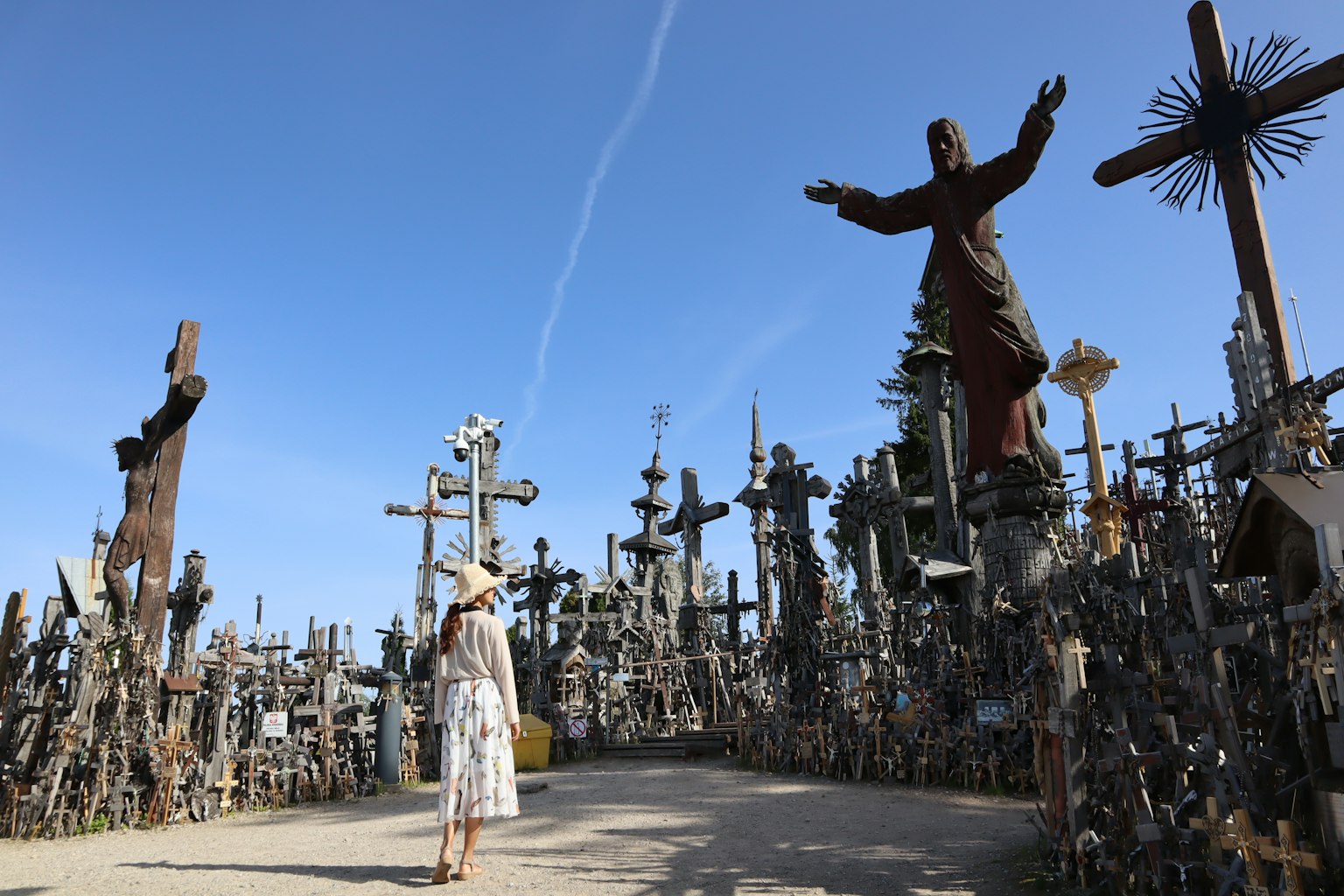 Mujer de pie en un sitio religioso rodeado de numerosas cruces y una gran estatua bajo un cielo azul