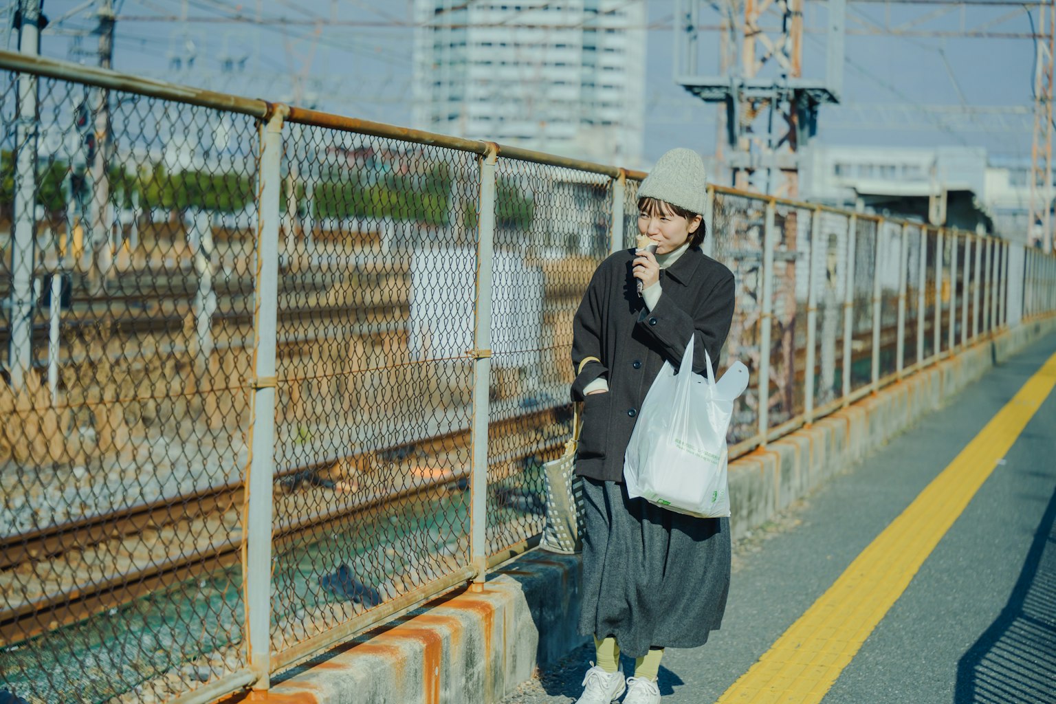 Young woman standing on a train platform smoking a cigarette wearing a black coat and gray skirt holding a white bag