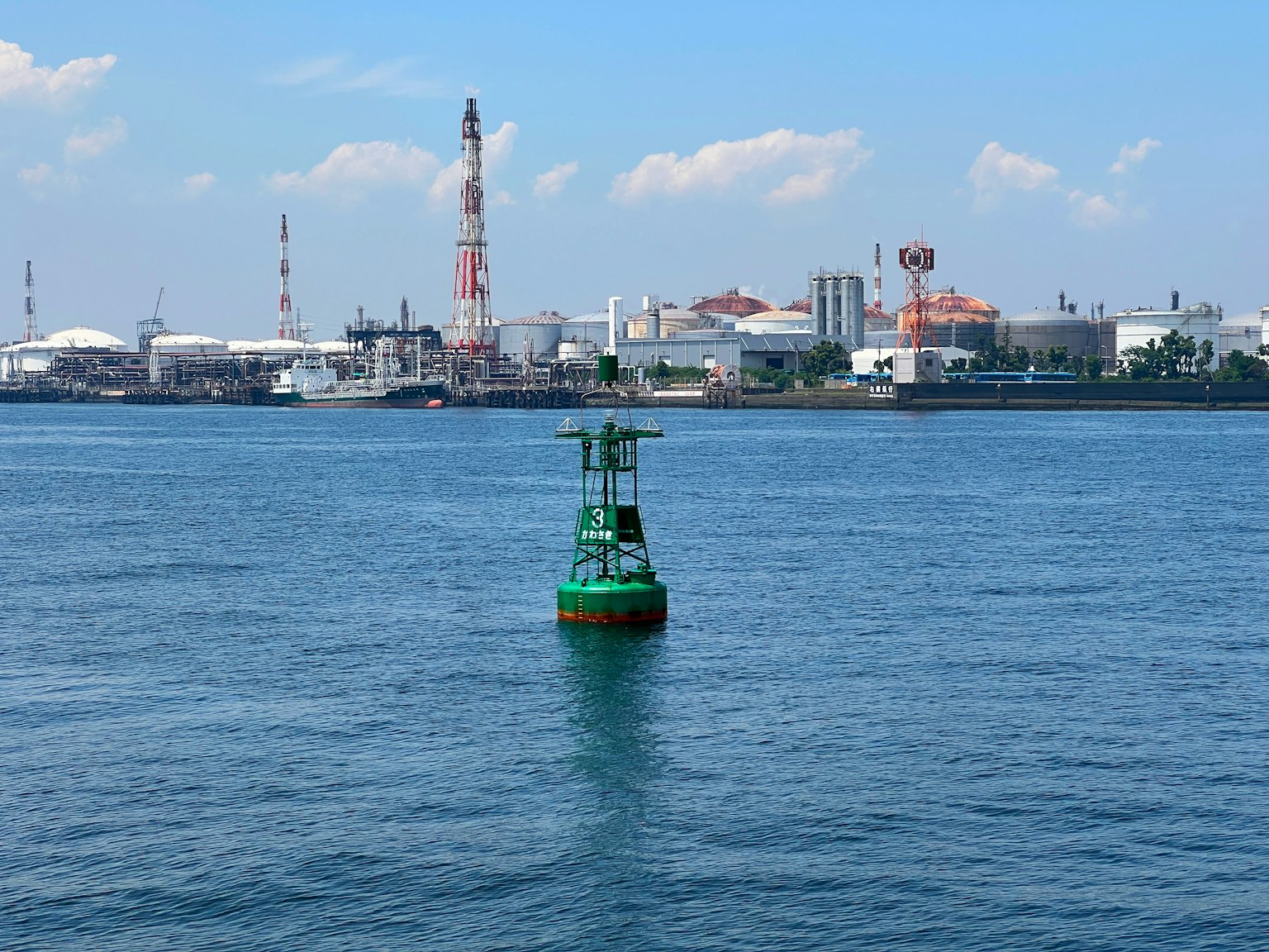 Una boya verde flotando en el agua con una zona industrial al fondo