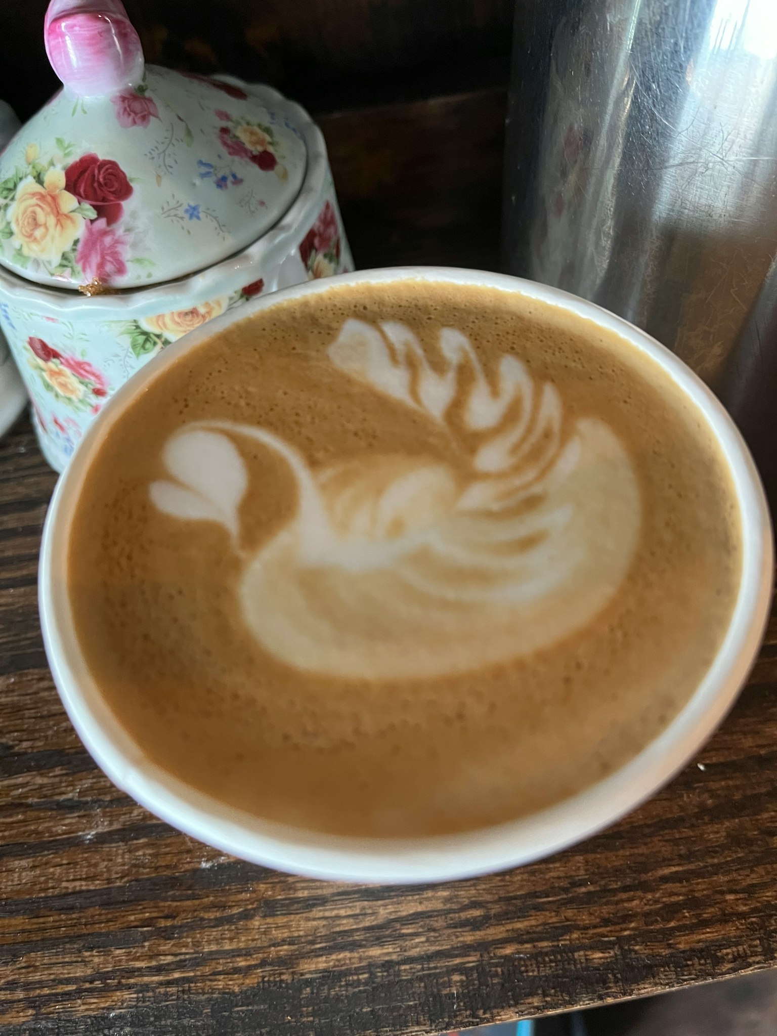 Latte art of a swan in a coffee cup with a floral lidded container nearby