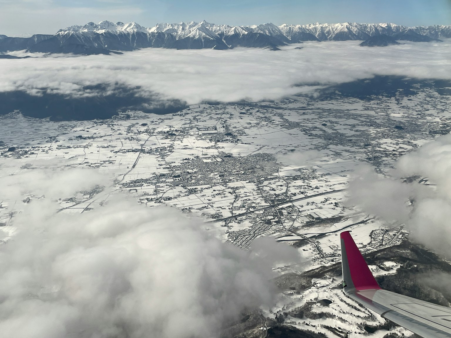 Flügel eines Flugzeugs über einer verschneiten Landschaft mit Bergen im Hintergrund