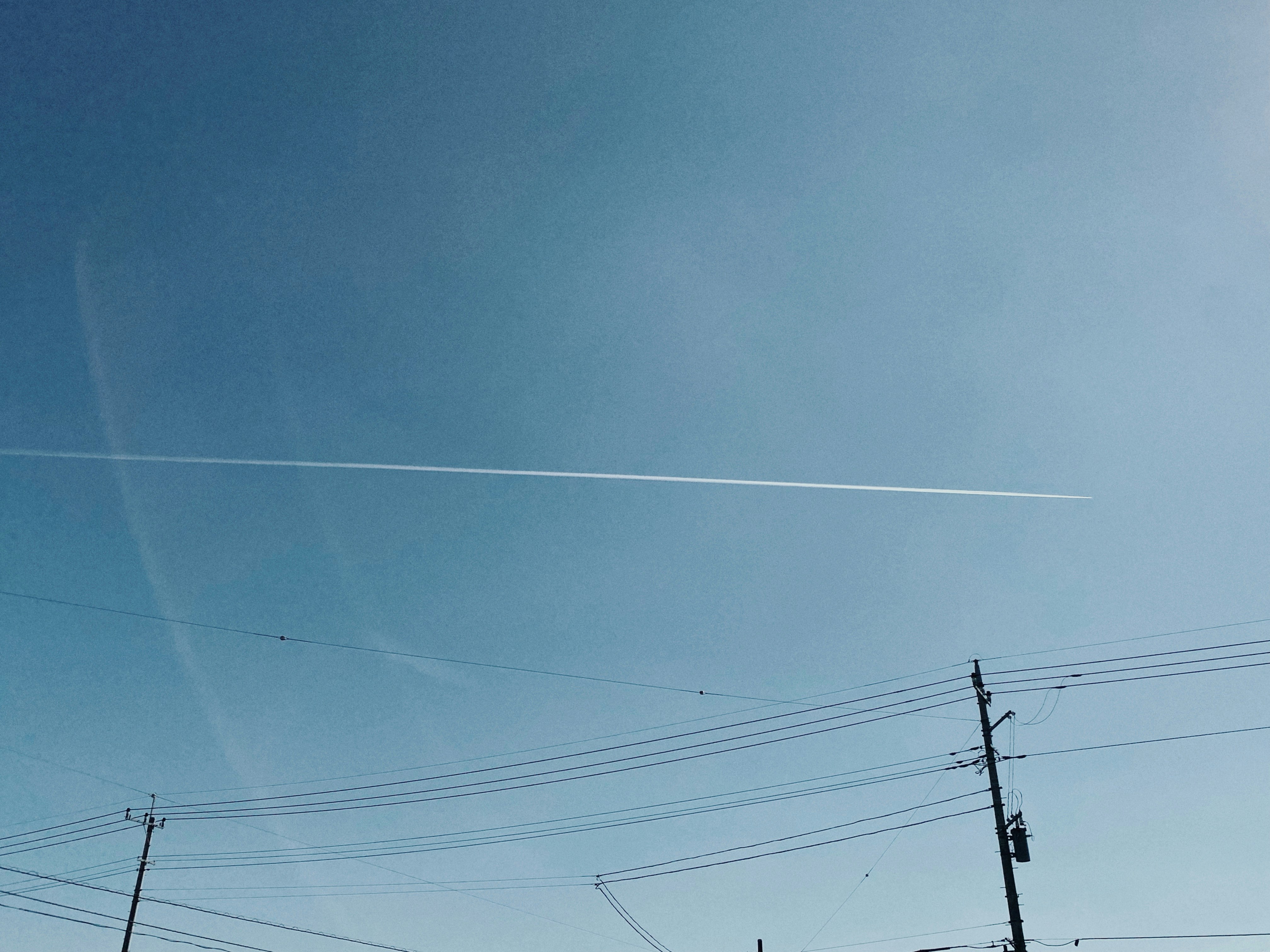 青空に伸びる飛行機雲と電線のある空の風景